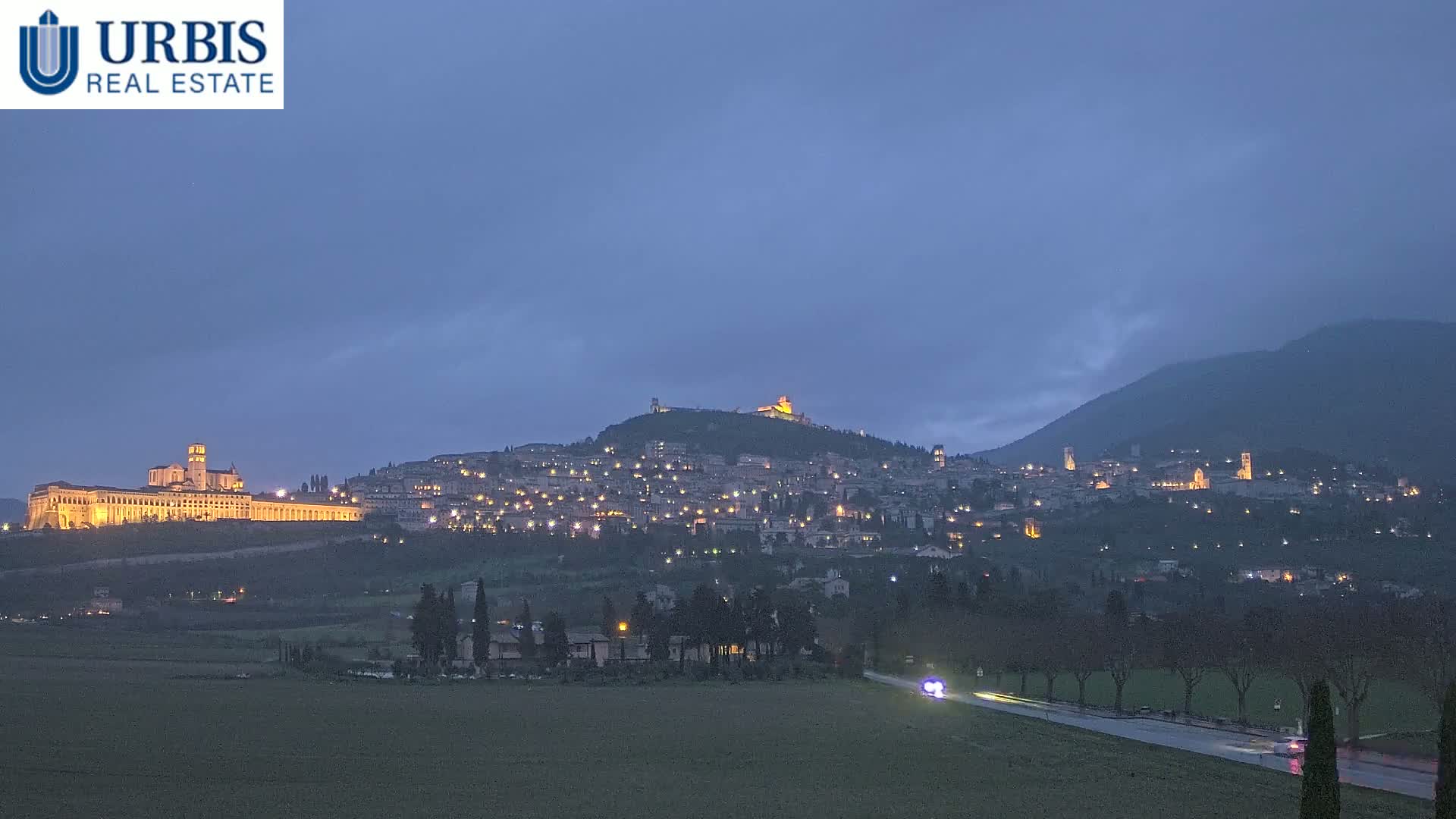 A hilltop town, illuminated at night, is seen from a distance under a dusky, overcast sky, with a road and a flat field in the foreground.