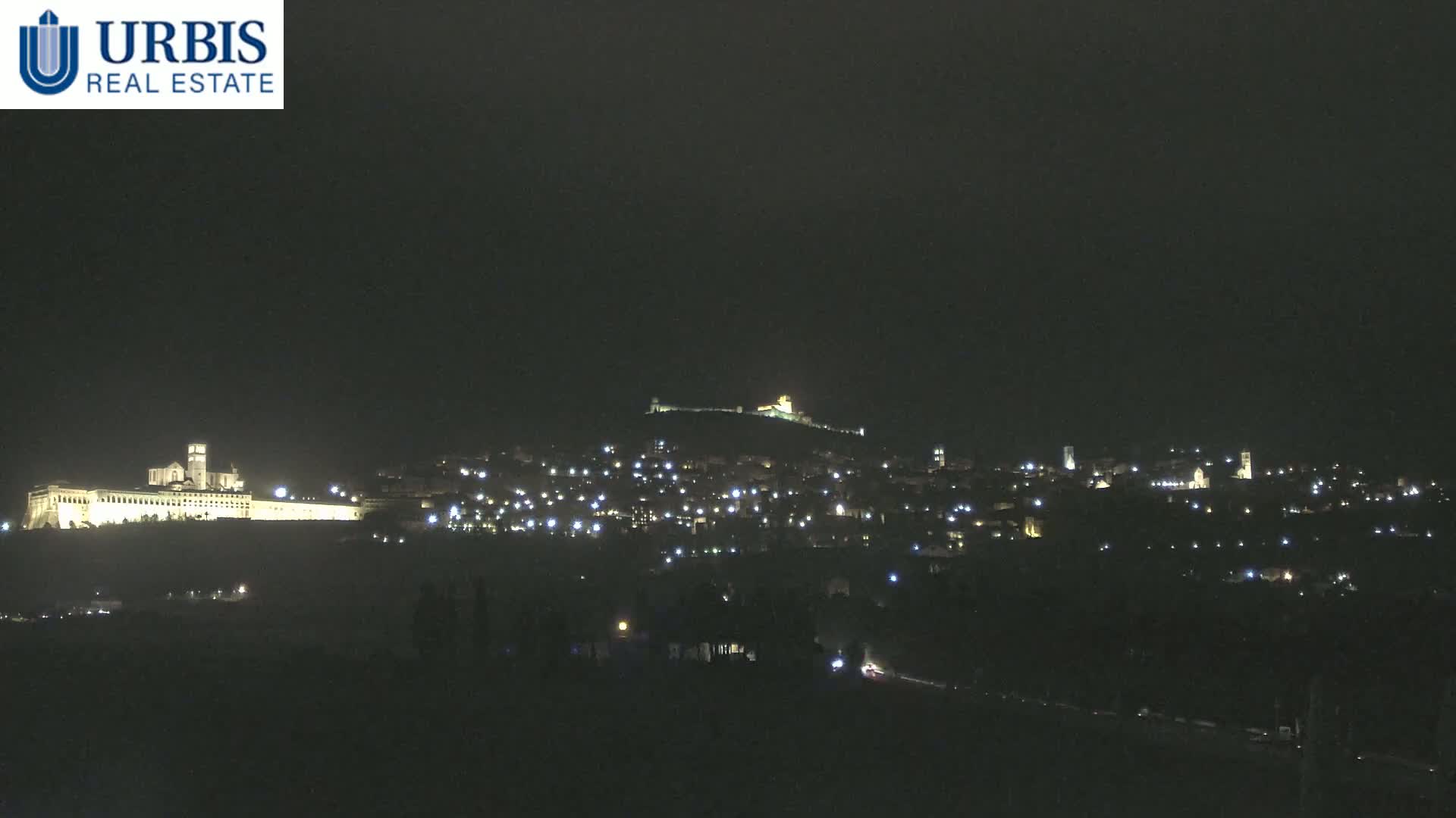 A nighttime view of a city on a hill, illuminated by numerous lights, with a large, brightly lit building at the base of the hill.