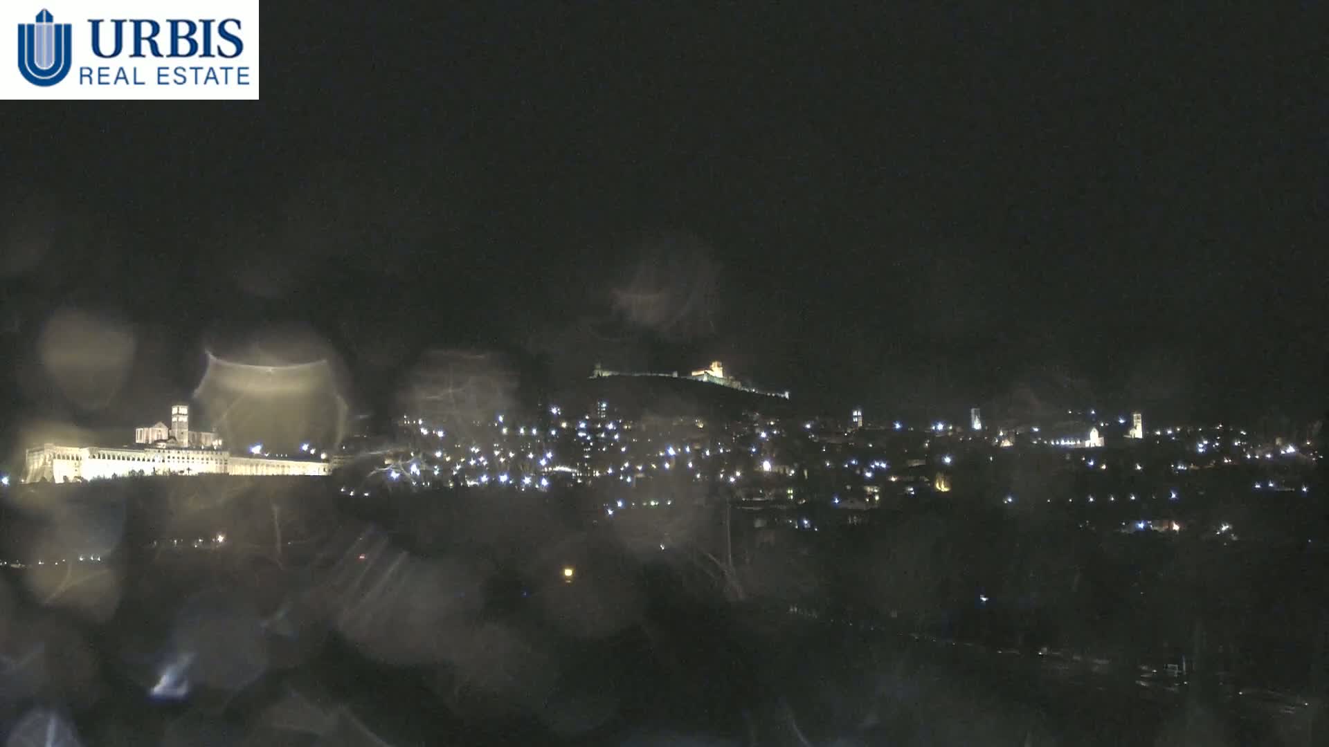 A nighttime view of a city illuminated by numerous lights, seen through a rain-covered window, with a hilltop structure in the distance.