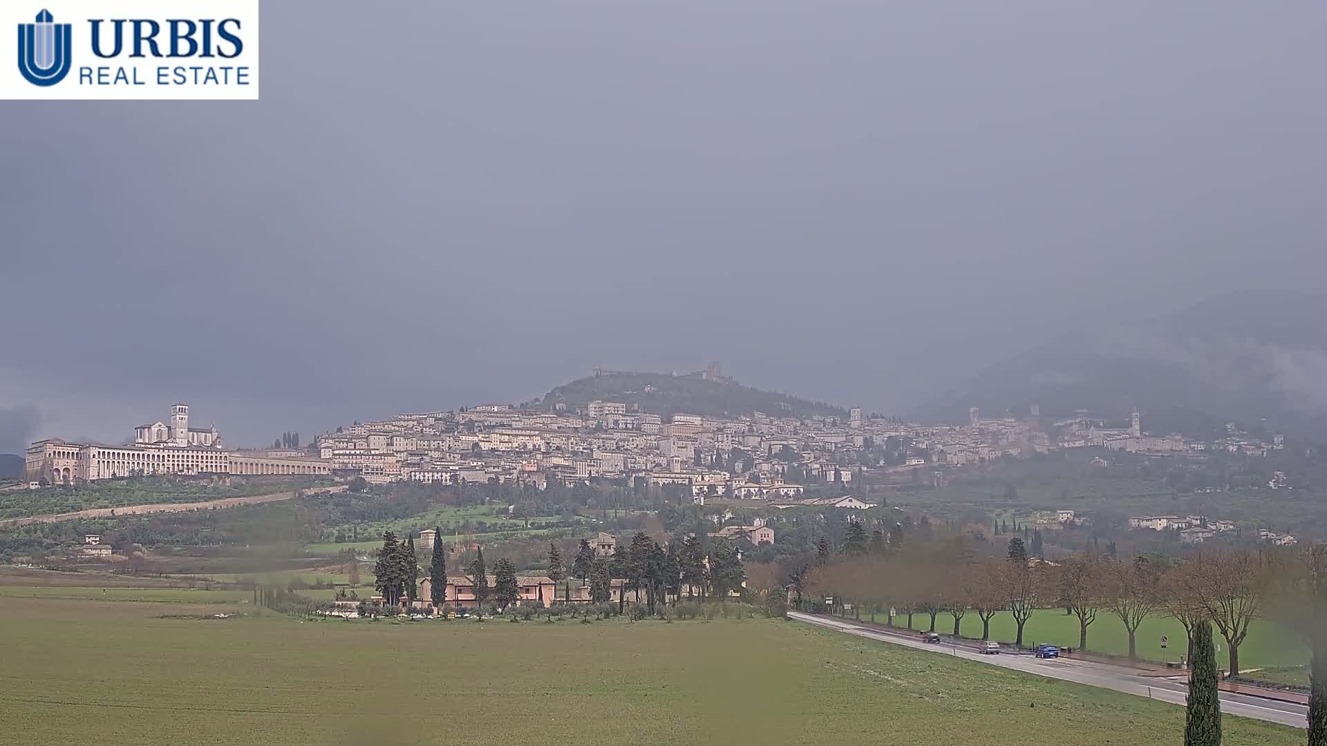 A hazy, overcast day reveals a large town nestled on a hill, with a long, low building at its base, and a flat, grassy area in the foreground.