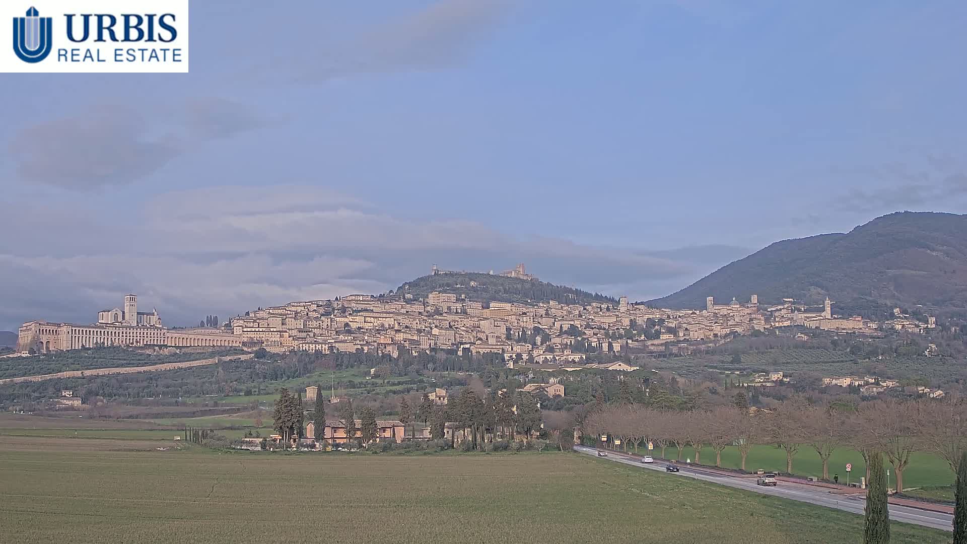 A large hilltop town with many tan buildings, a long basilica, and surrounding green fields is visible under a mostly clear, pale blue sky.