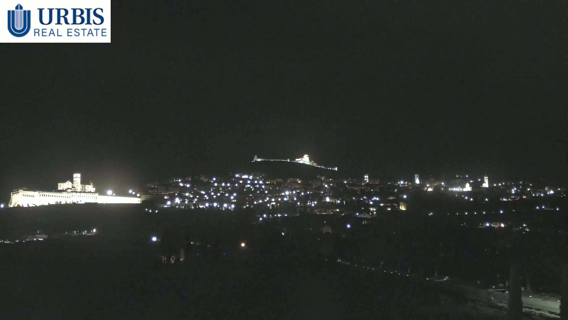 A nighttime view shows a hilltop town illuminated by scattered lights, with a large, brightly lit structure on a lower hill in the foreground.