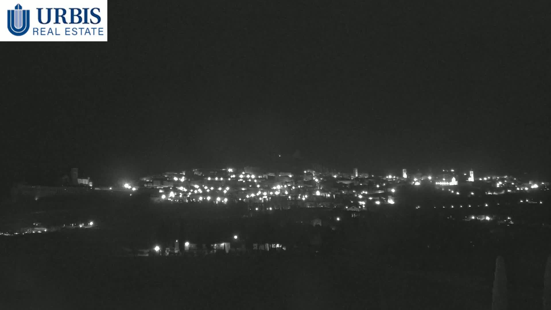 A nighttime long shot shows a small town illuminated by many scattered lights on a hill under a dark, clear sky.