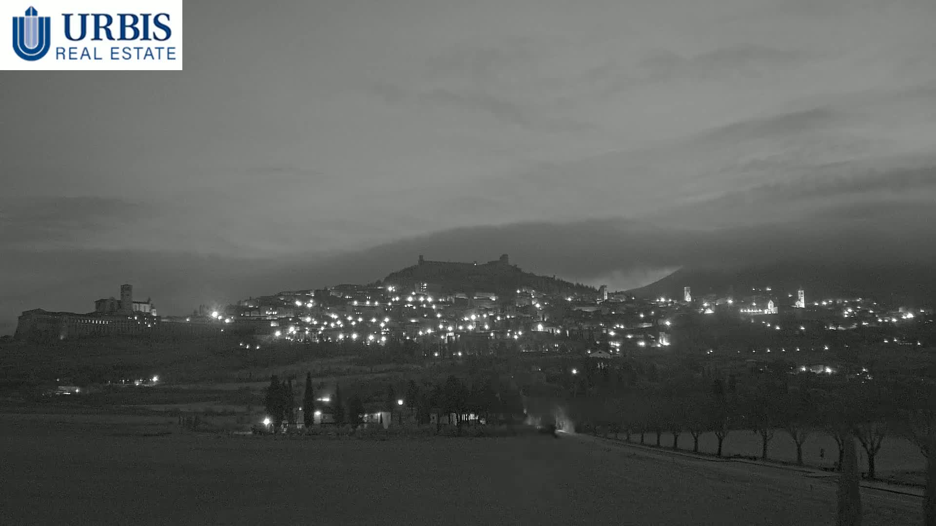 A hilltop town, illuminated at night under a cloudy, dusky sky.