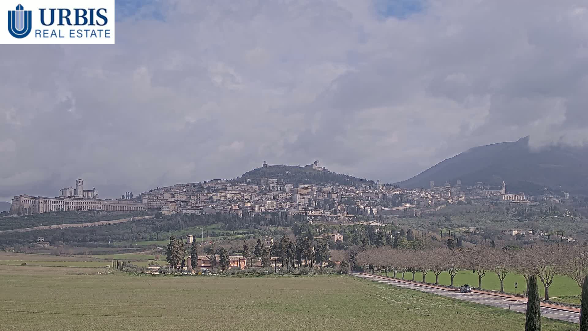 A hilltop town with a large basilica and a long wall, under a cloudy sky, is seen from across a flat field with a road running through it.
