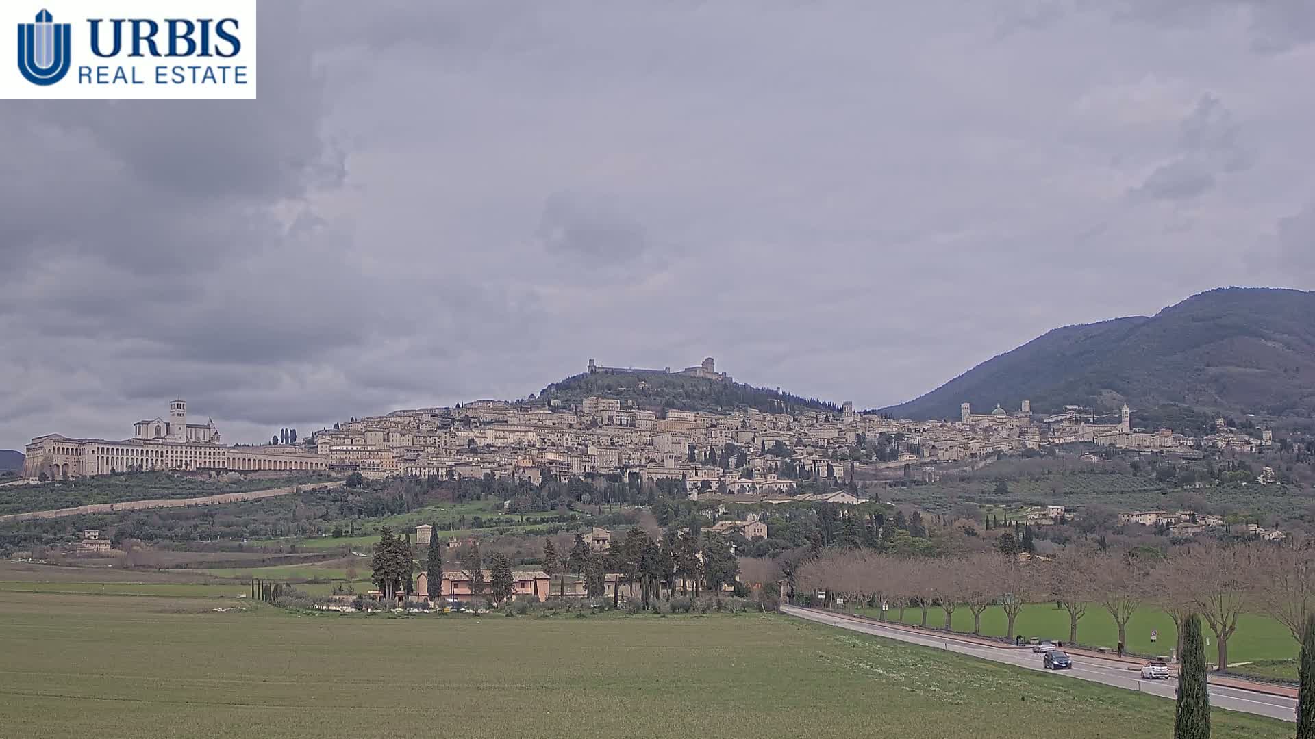 A sprawling hilltop town, under a cloudy sky, is viewed from a distance across a flat, grassy plain with a road and sparse trees.