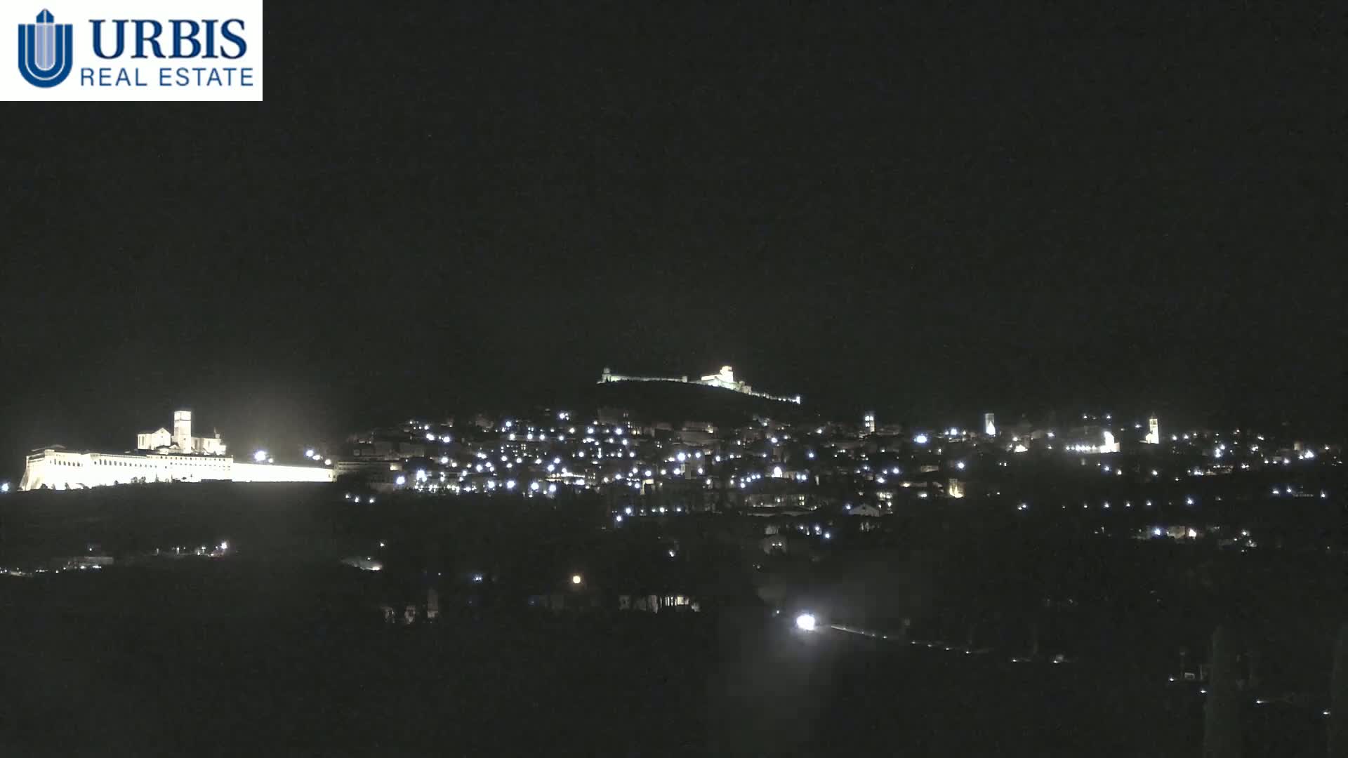 A nighttime long shot shows a hilltop town with numerous illuminated buildings and a large, brightly lit structure at the base of the hill.