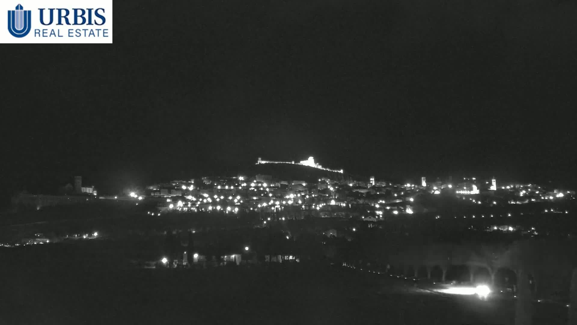 A nighttime long shot shows a hilltop town illuminated by its lights under a clear, dark sky.
