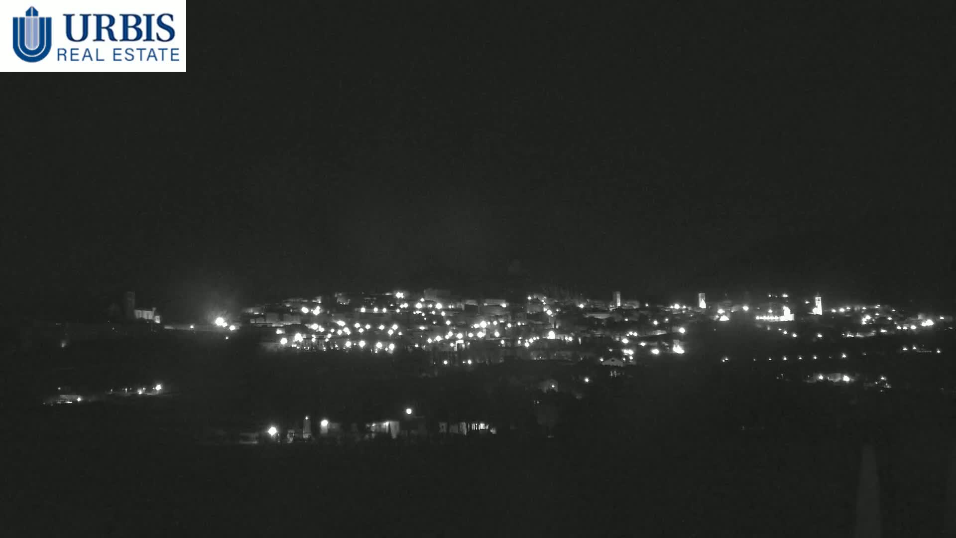 A nighttime long shot shows a small town nestled on a hill, illuminated by scattered lights under a dark, clear sky.