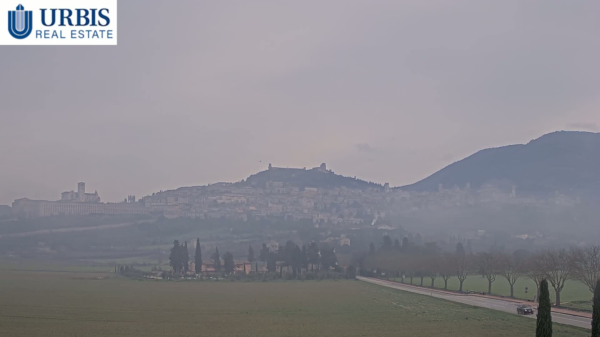A hazy view of a town nestled on a hill, with a larger structure at its base, and a mountain in the background under a gray sky.