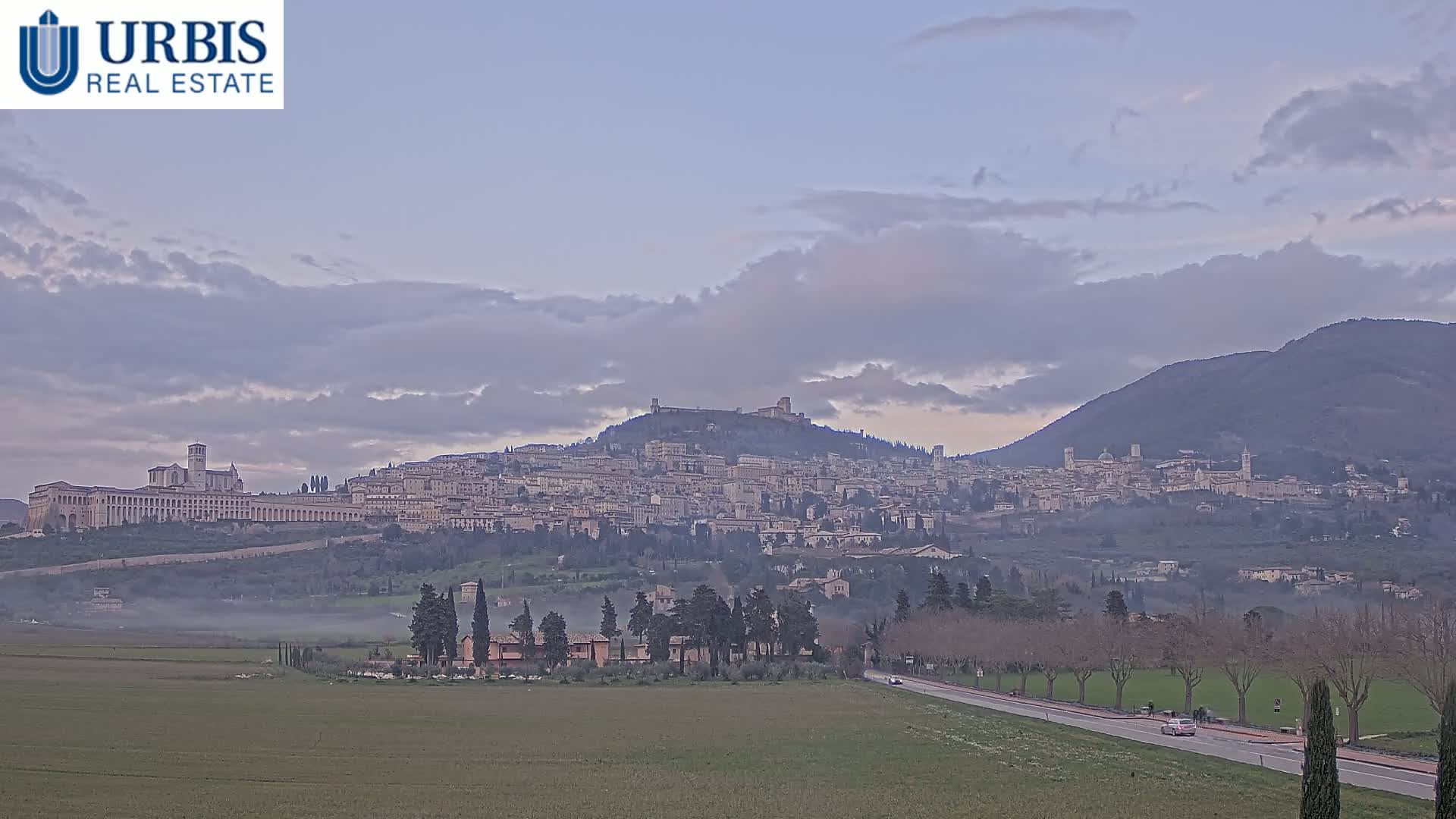 A hazy, overcast sky hangs over a hilltop town, nestled between rolling hills and a valley shrouded in low-lying fog.