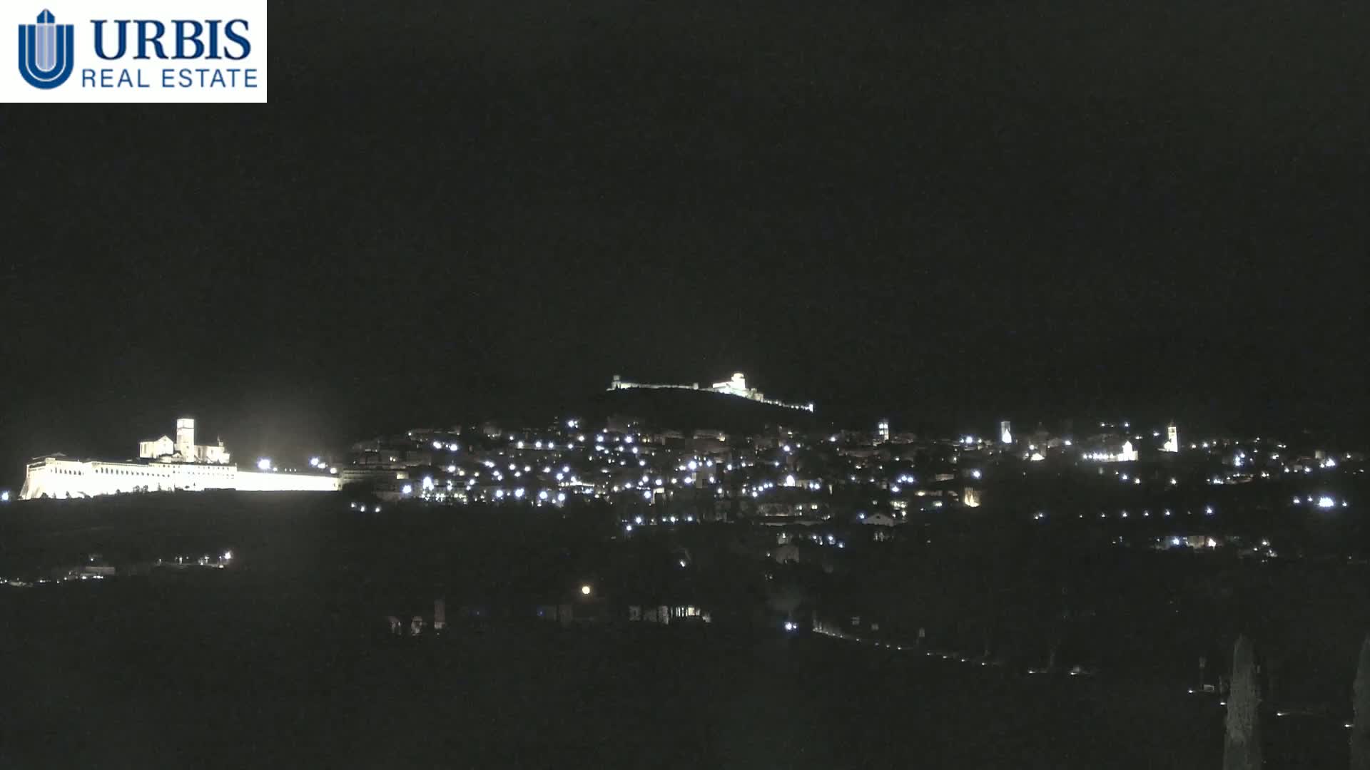 A nighttime view of a town illuminated by lights, situated on a hill with a larger, brightly lit structure on a hill in the background.