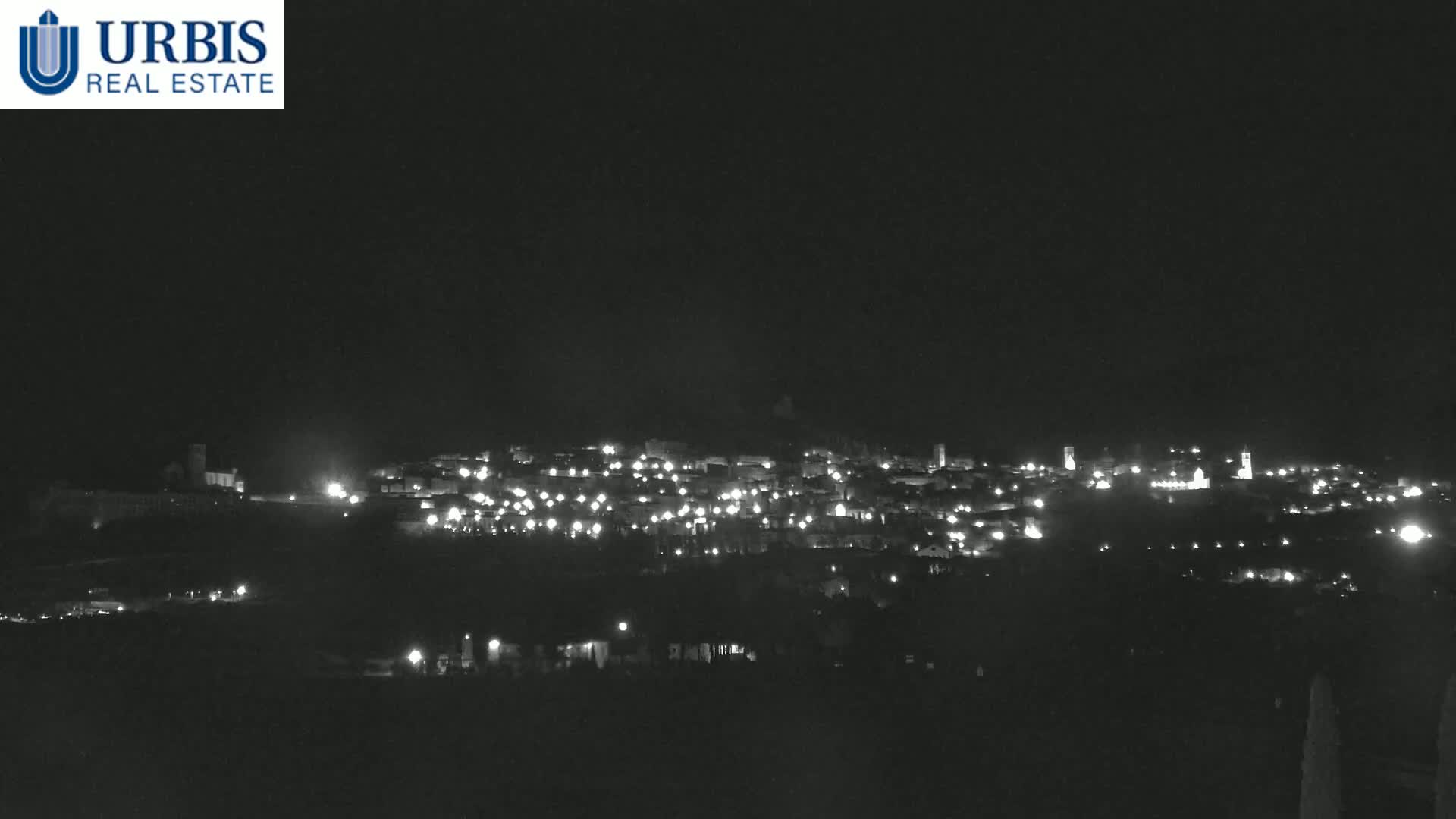 A nighttime long shot shows a small town illuminated by scattered lights on a hill under a dark, clear sky.