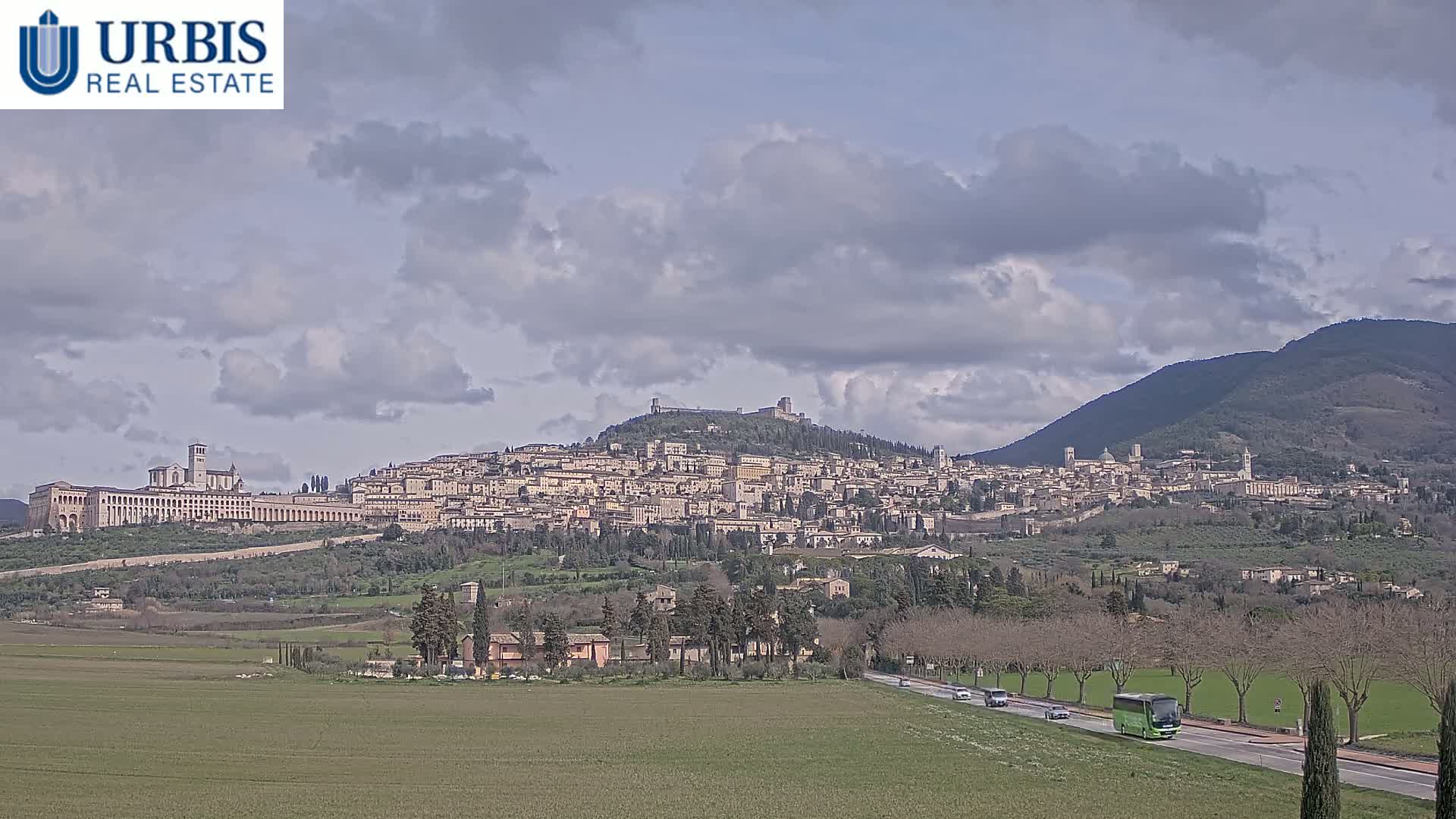 A large town sits atop a hill under a partly cloudy sky, with a road and green fields in the foreground.