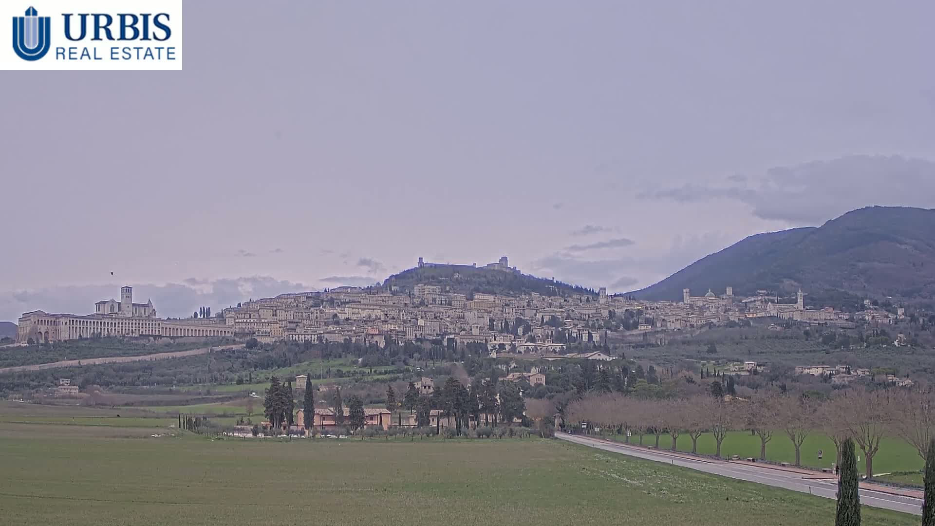 A sprawling hilltop town with numerous buildings, including a large basilica, is visible under an overcast sky, set against a backdrop of rolling hills and a mountain.