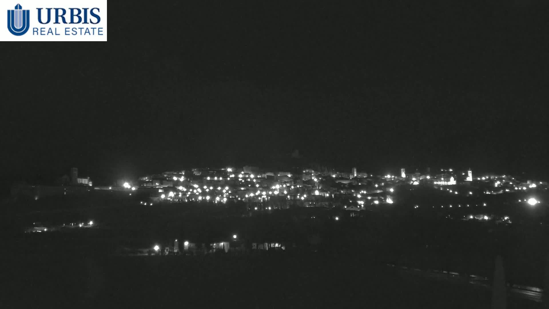 A nighttime long shot shows a town illuminated by many small lights spread across a hillside under a dark, clear sky.