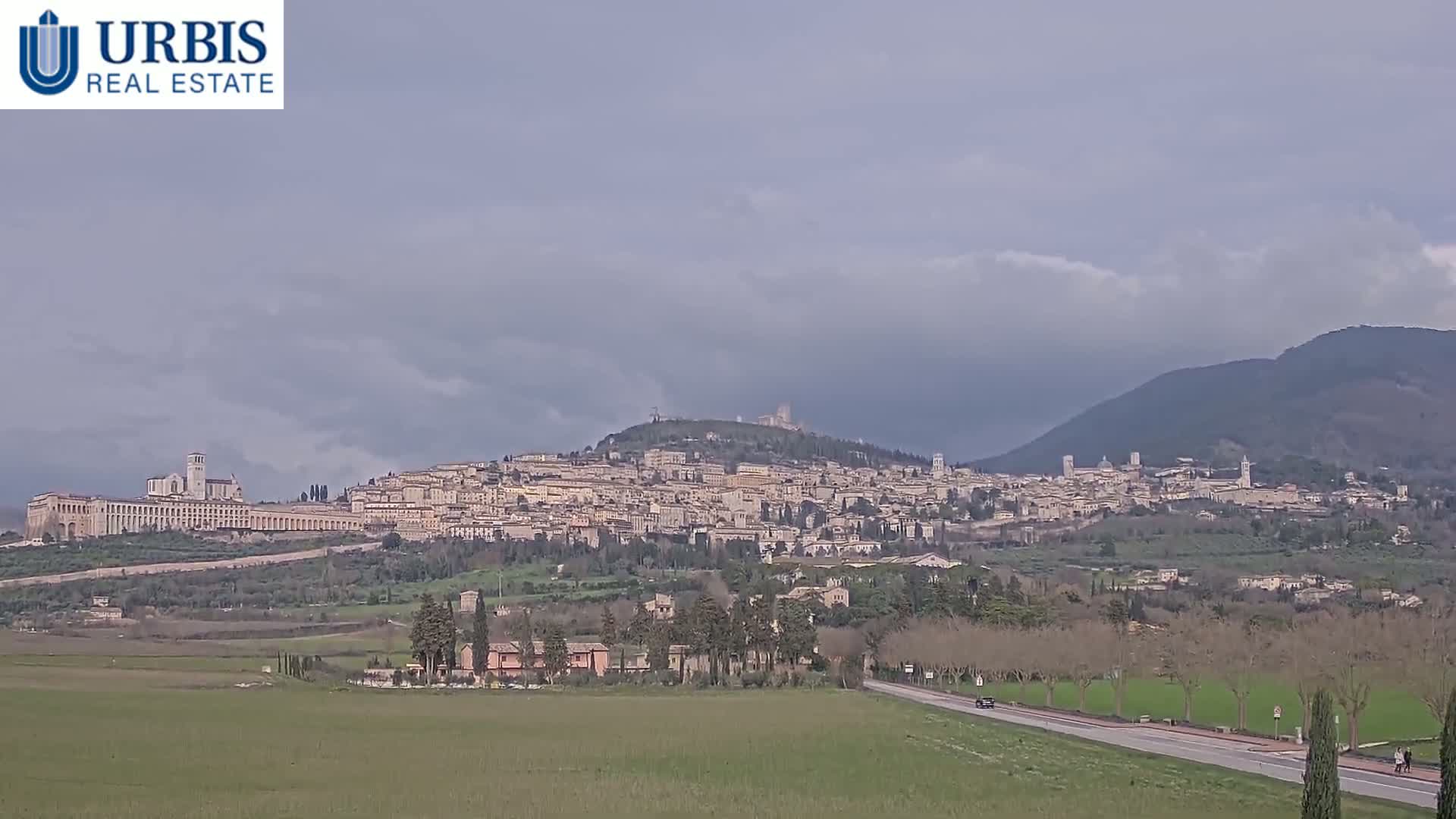 A large hill town, under a cloudy sky, is visible from a distance across a flat, grassy plain, with a road and sparse trees in the foreground.