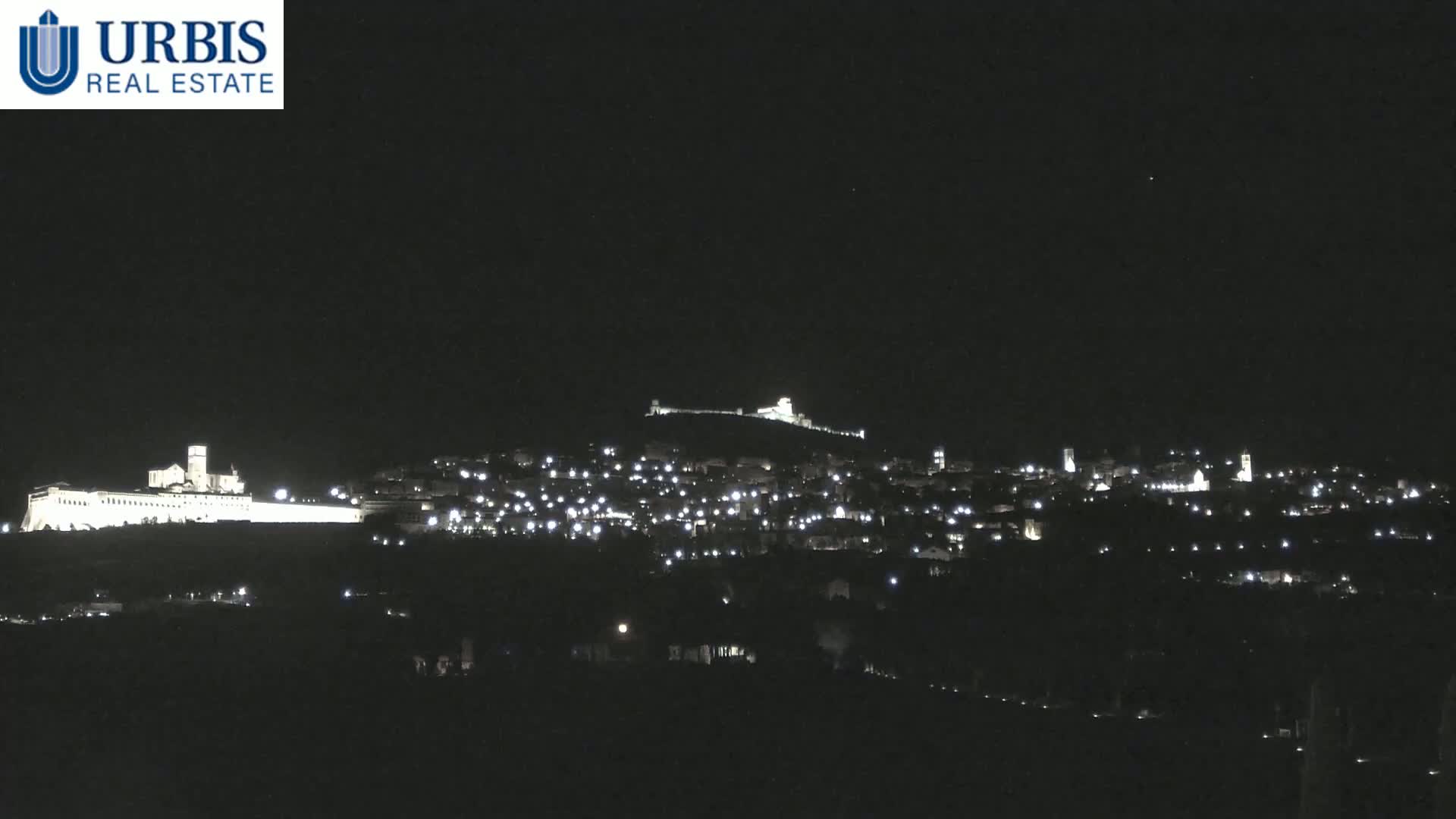 A nighttime view of a town nestled on a hill, illuminated by scattered lights, with a larger, brighter structure situated on a higher hill in the background under a clear, dark sky.