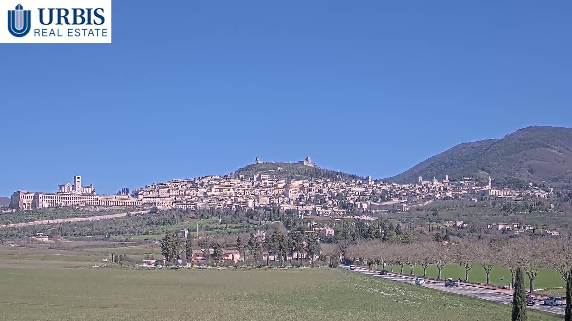 A large town sprawls across a hillside under a clear blue sky, with a road and flat green fields in the foreground.