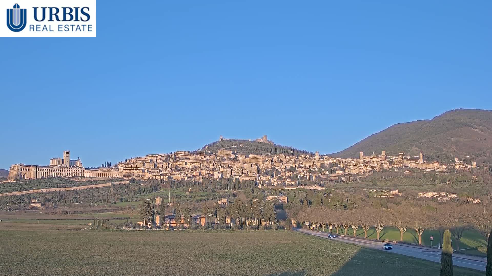A large town sits atop a hill under a clear blue sky, with a long, flat building and a mountain visible in the background.