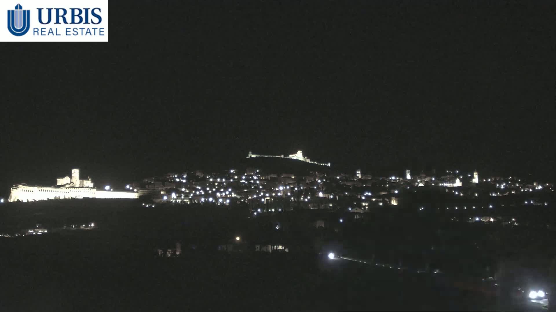 A nighttime view of a hilltop town illuminated by lights, featuring a large, brightly lit building on a lower hill in the foreground under a clear, dark sky.