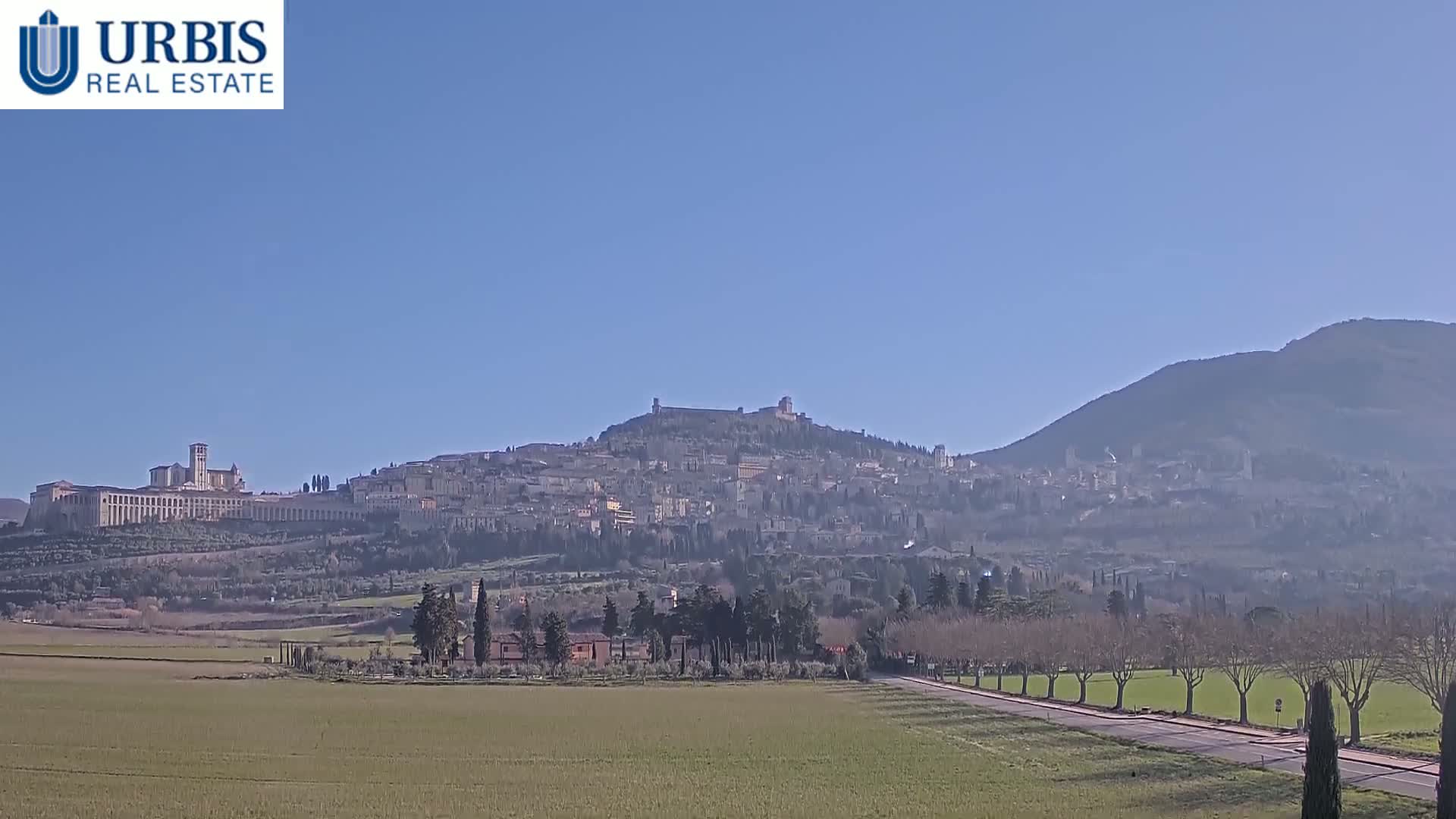 A distant view of a hilltop town, under a clear blue sky, is seen from across a flat field with a road leading toward it.
