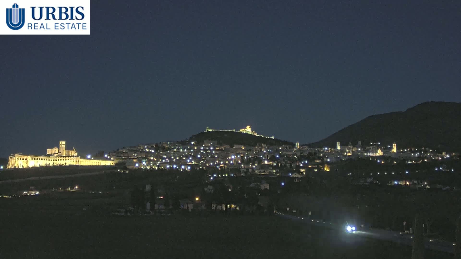 A nighttime view of a hilltop town, illuminated by lights, with a large, brightly lit building complex on a lower hill in the background under a clear, dark sky.