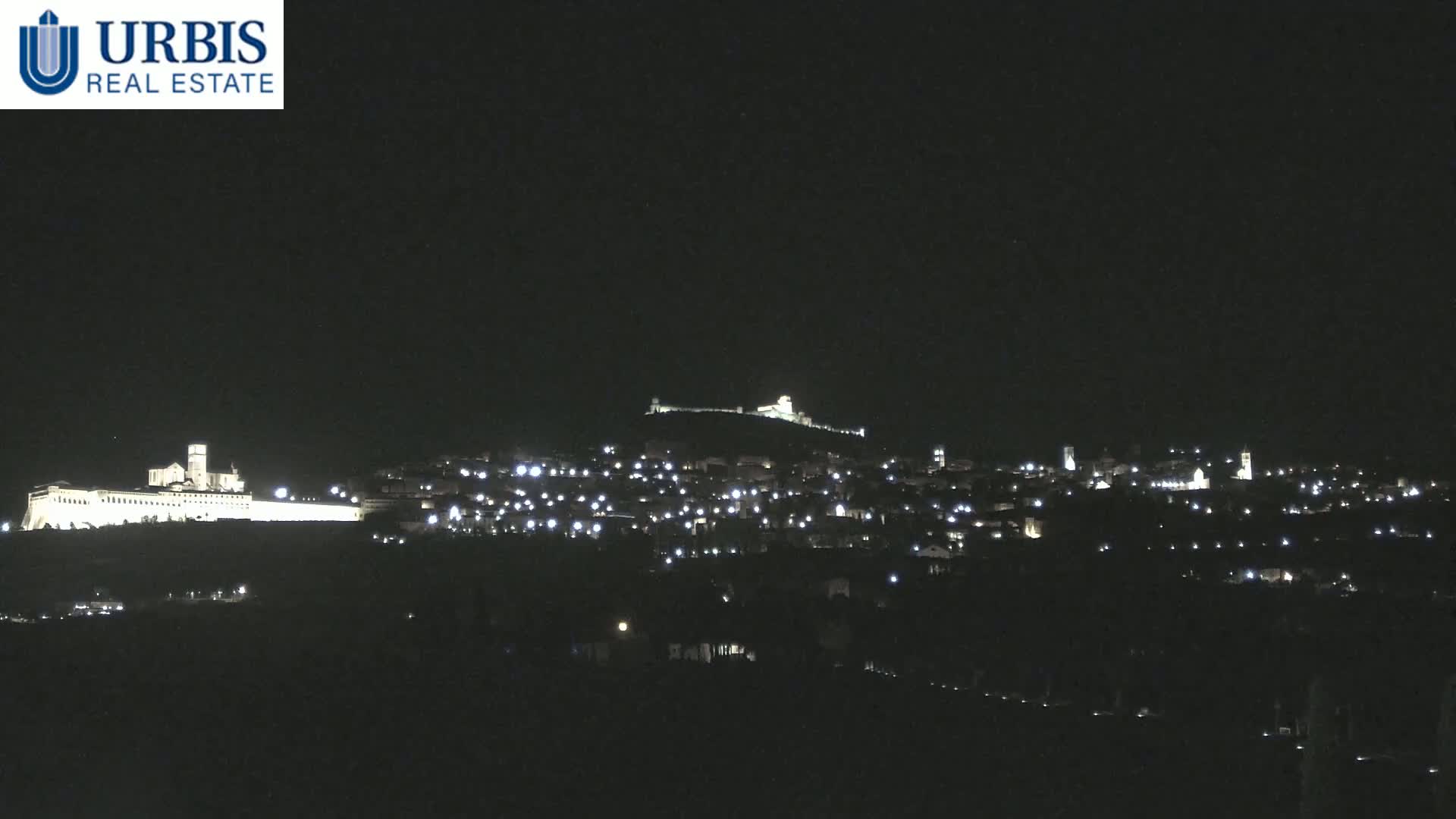 A nighttime view of a town nestled on a hill, illuminated by scattered lights, with a large, brightly lit building on a lower hill in the foreground.