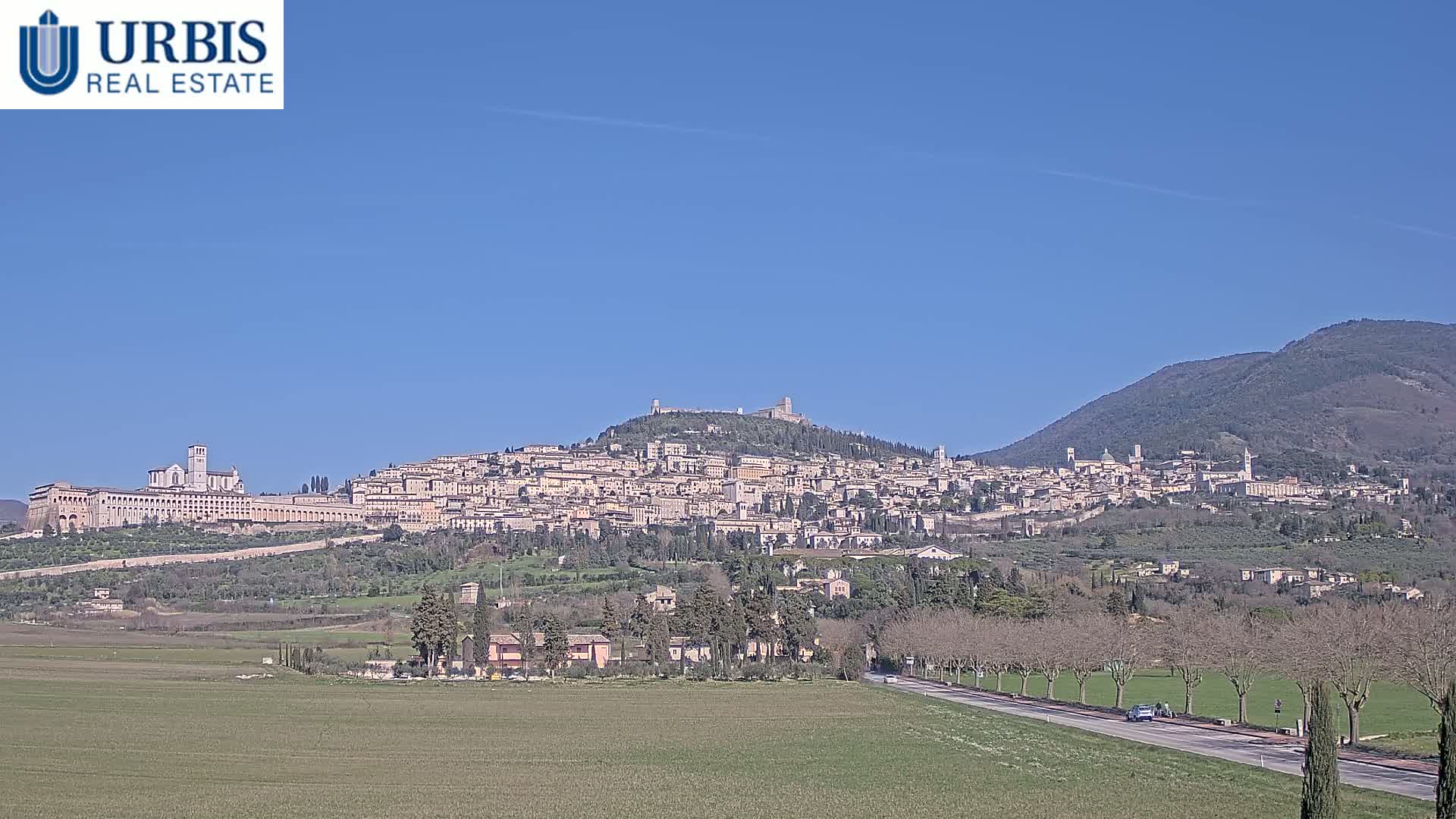 A hilltop town with many buildings, including a large church, is seen in the distance on a clear, sunny day, with a flat green field in the foreground and mountains in the background.