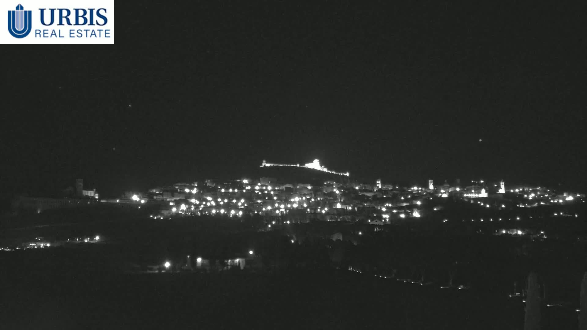 A nighttime, long shot of a town nestled on a hill, illuminated by scattered lights, under a clear, dark sky.