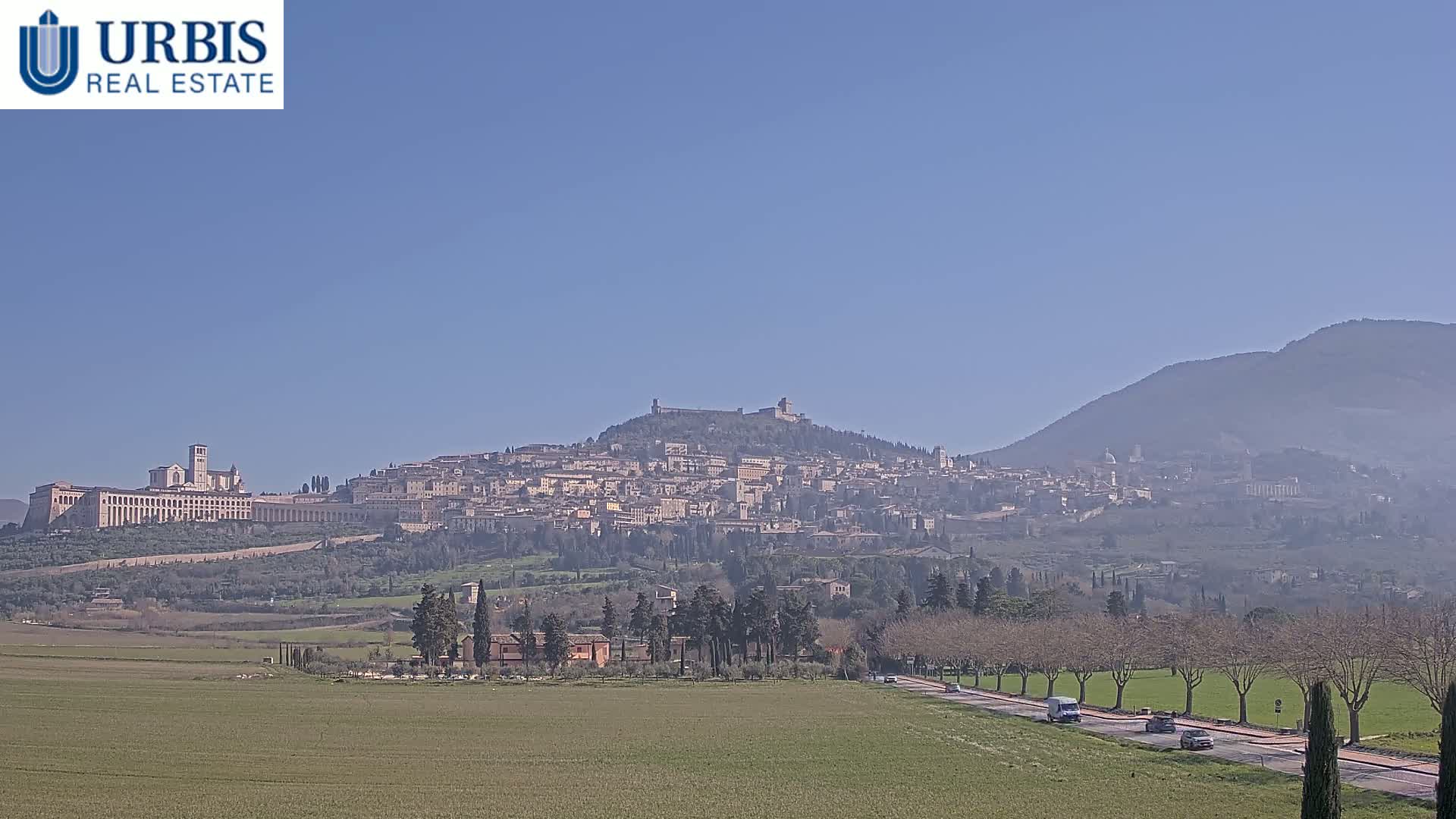 A hilltop town, possibly Assisi, stretches across a mountain under a clear blue sky, with a road and a field in the foreground.