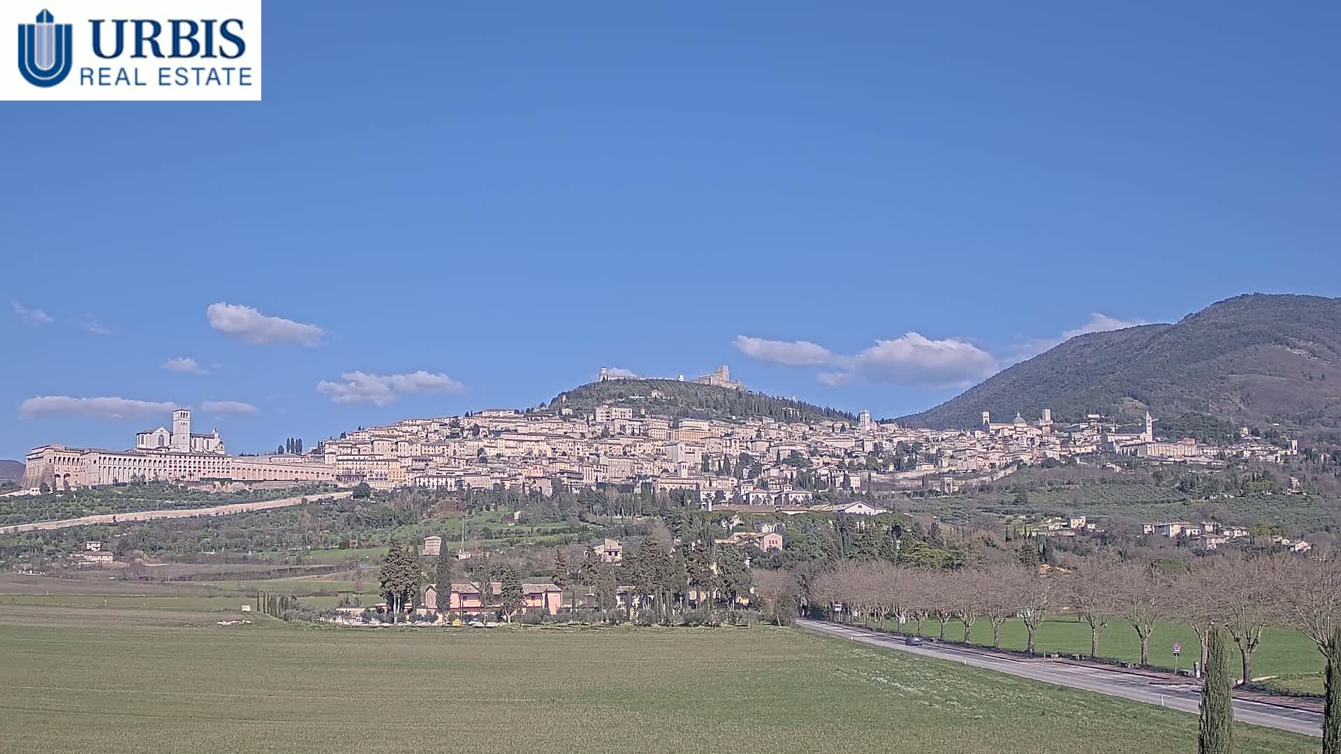 A hilltop town, under a mostly clear blue sky, stretches across a hillside with a large building prominently featured in the foreground.