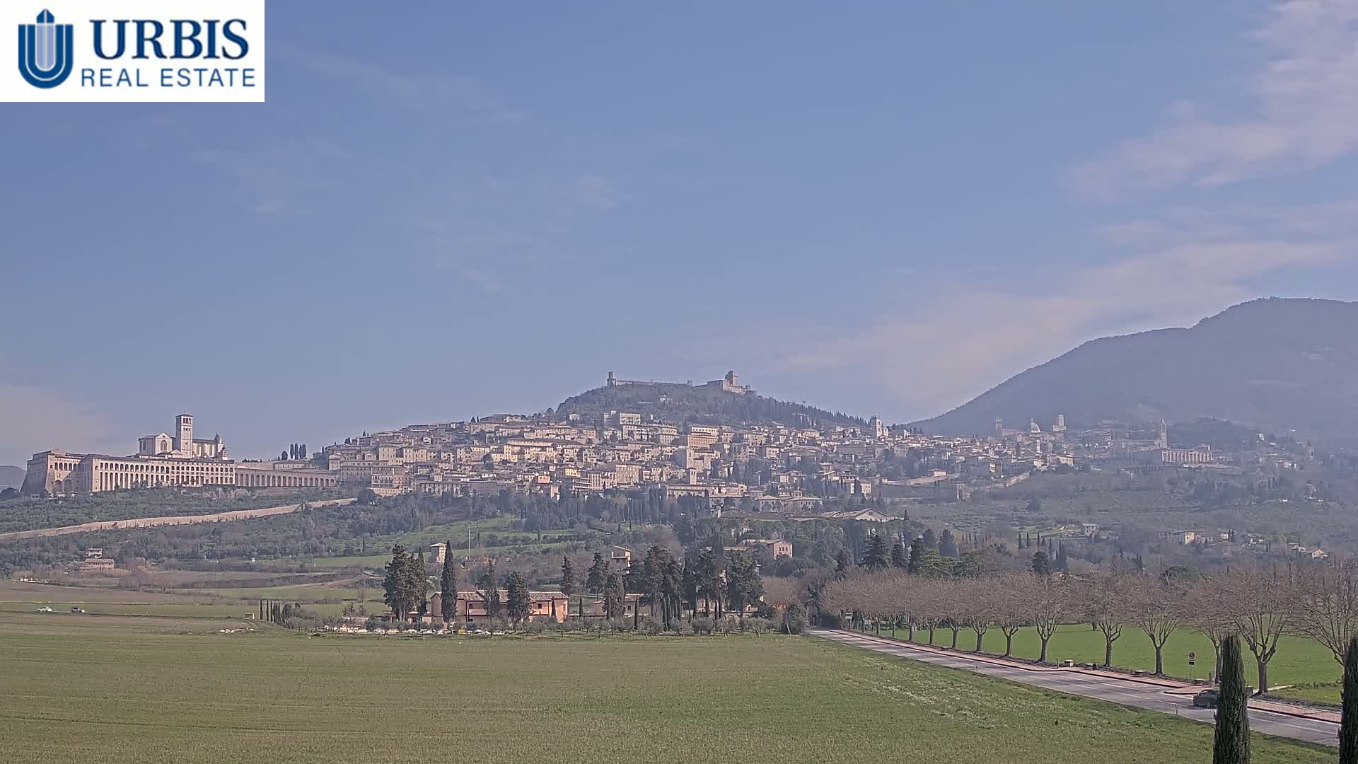 A hilltop town with numerous buildings, including a large church, is seen in the distance under a clear sky, with a road and fields in the foreground.