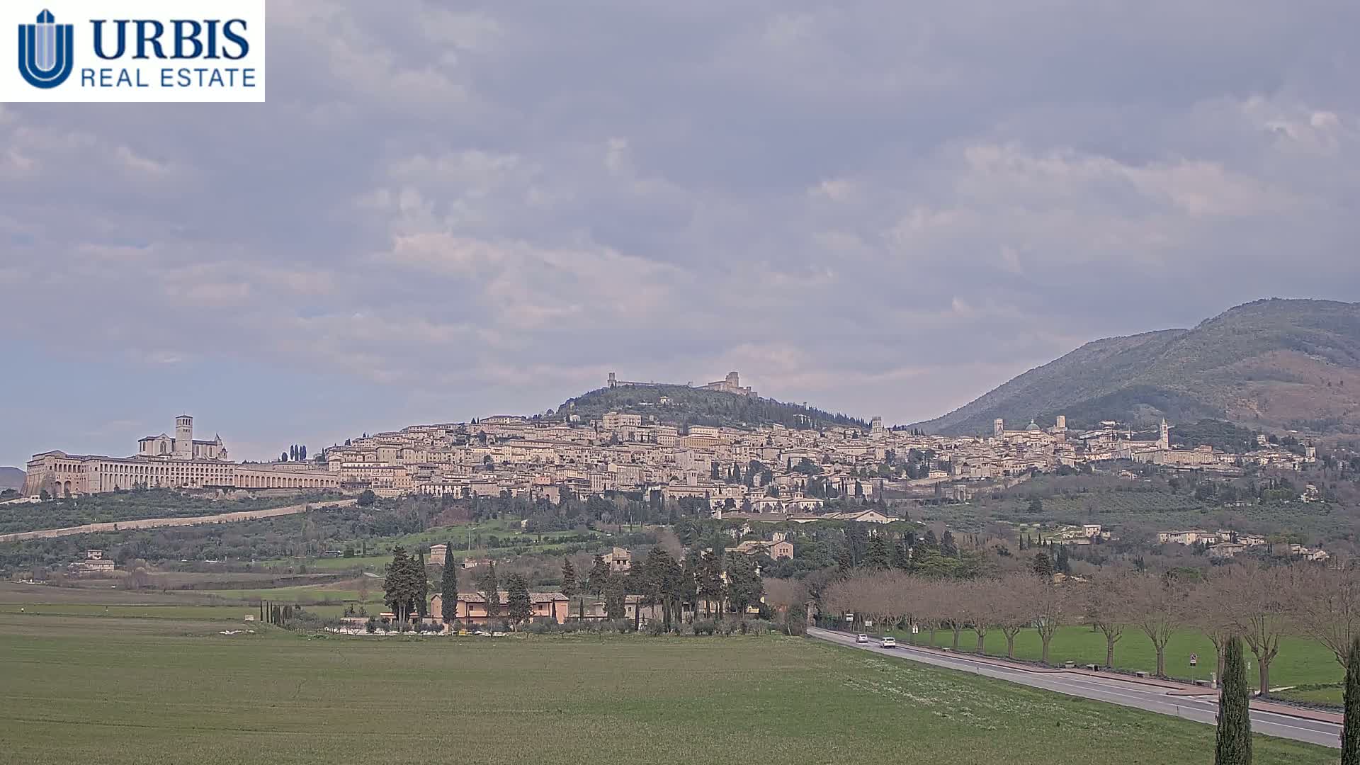A sprawling hilltop town, under a mostly cloudy sky, is viewed from a distance across a flat, grassy field and a road lined with bare trees.