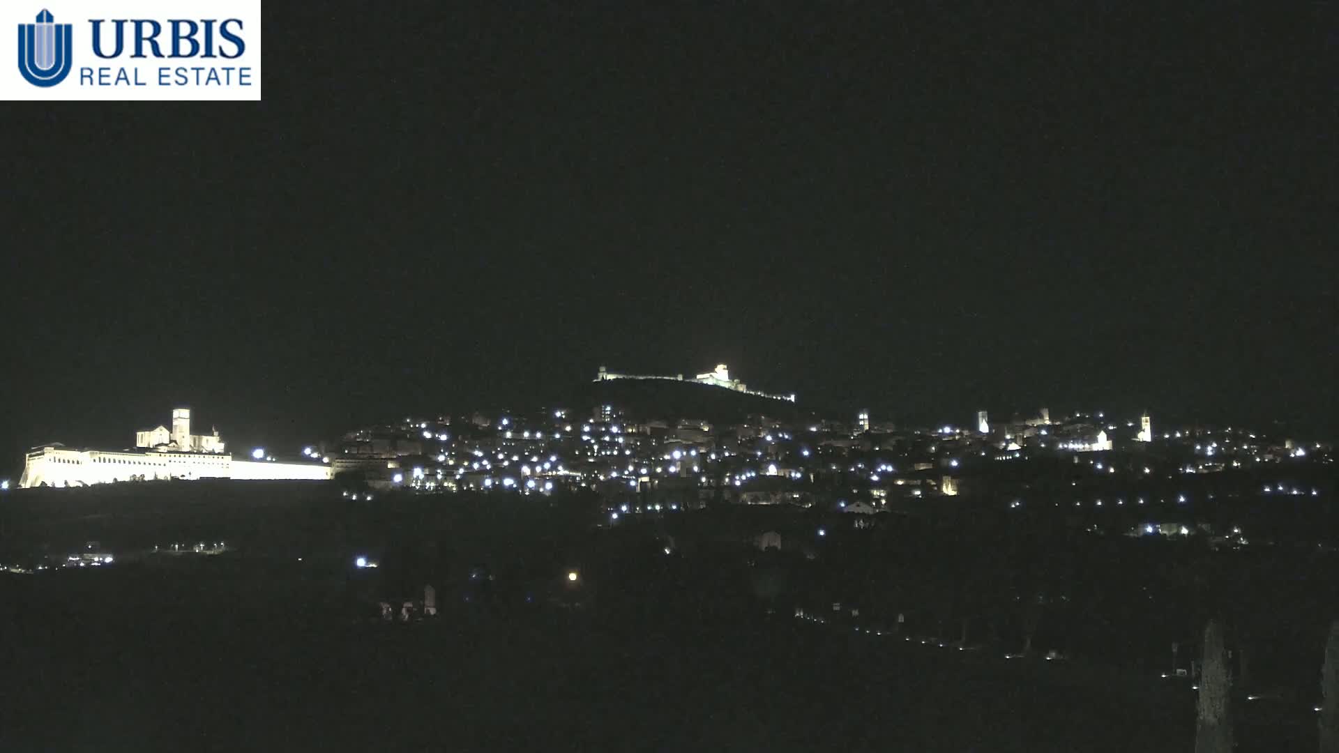 A nighttime long shot of a town nestled on a hill, illuminated by scattered lights, with a larger, brightly lit building complex situated on a lower hill in the foreground under a clear, dark sky.