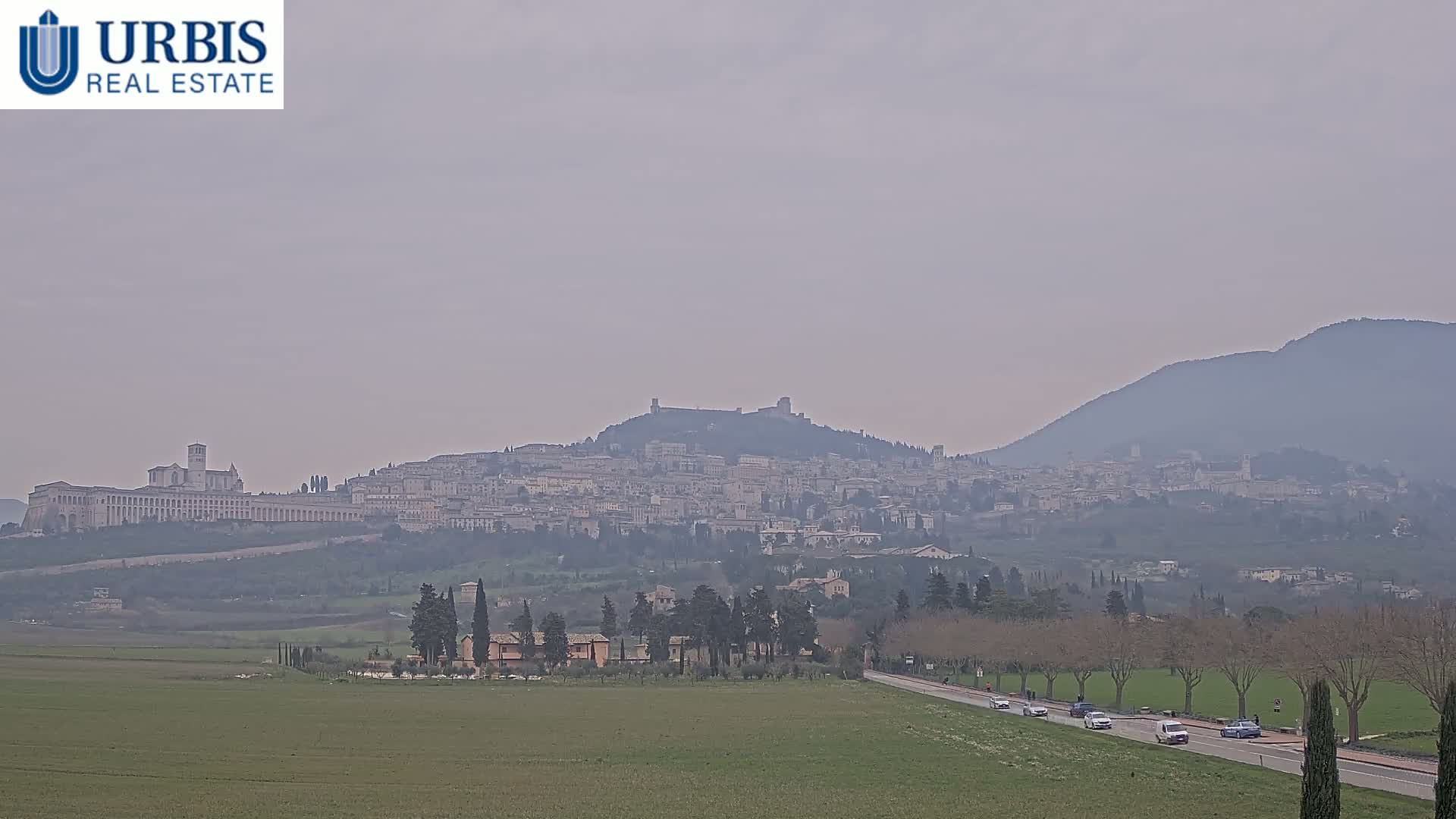 A hazy view shows a large hilltop town with a long, light-colored building at its base, all nestled between rolling green hills and a distant mountain range.
