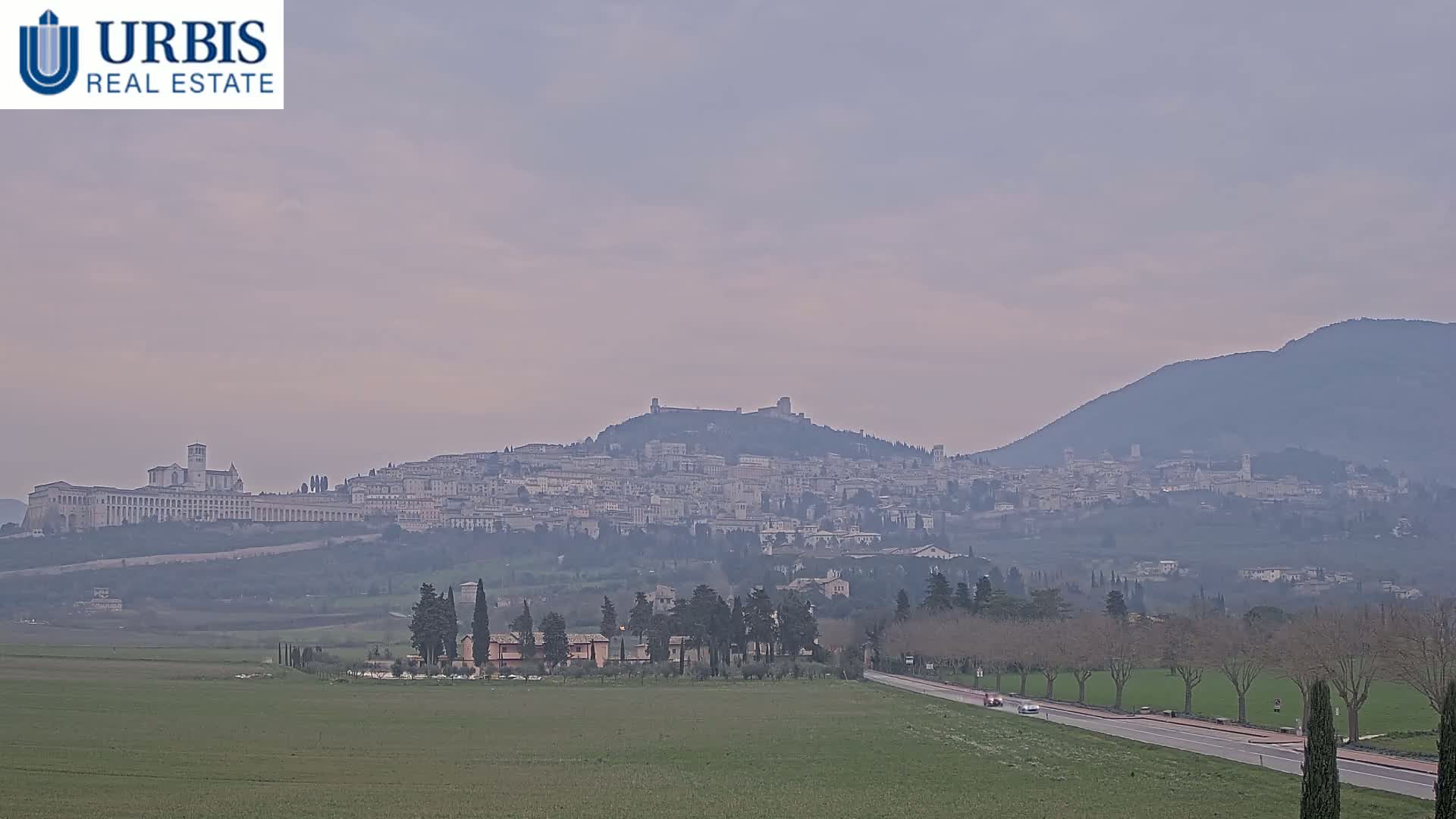 A hazy view shows a large town nestled on a hill, with a long basilica-like building extending across a portion of the hill, and mountains in the background.