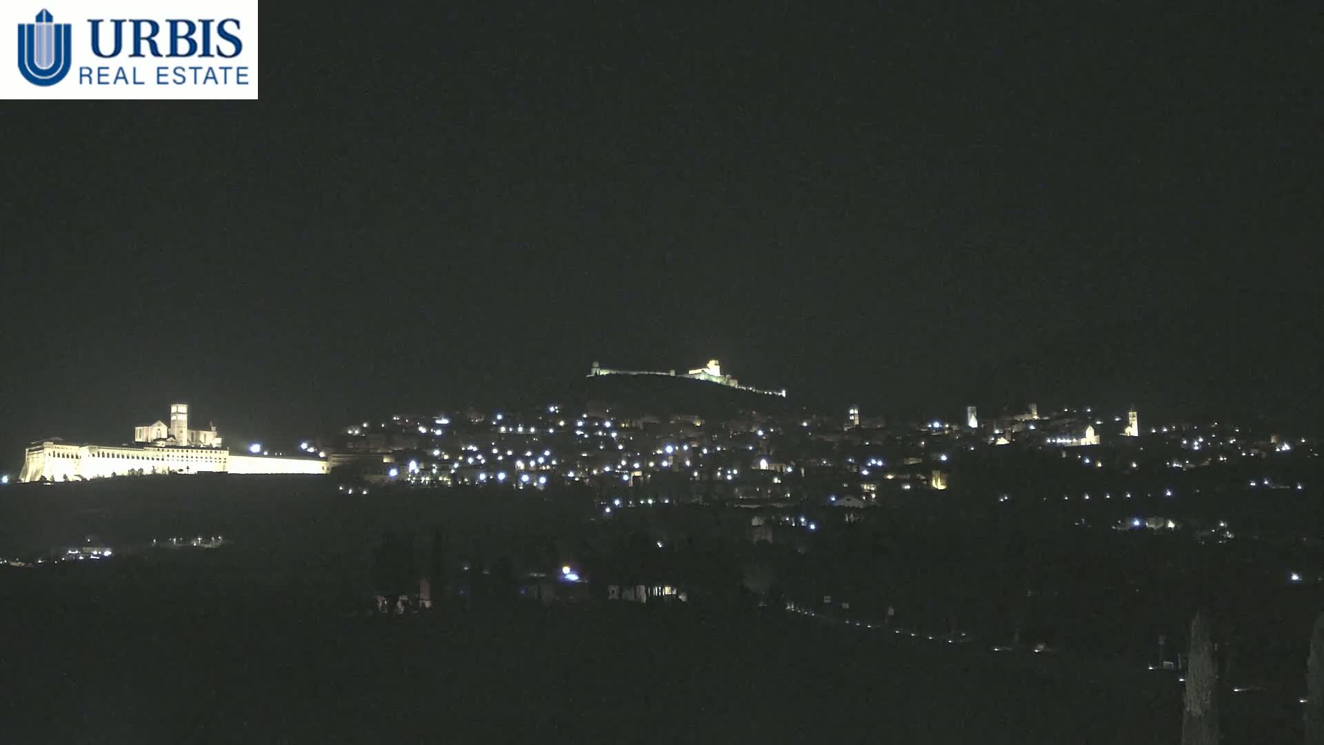 A nighttime view of a town illuminated by scattered lights on a hill, with a larger, brightly lit structure on a lower hill in the foreground.