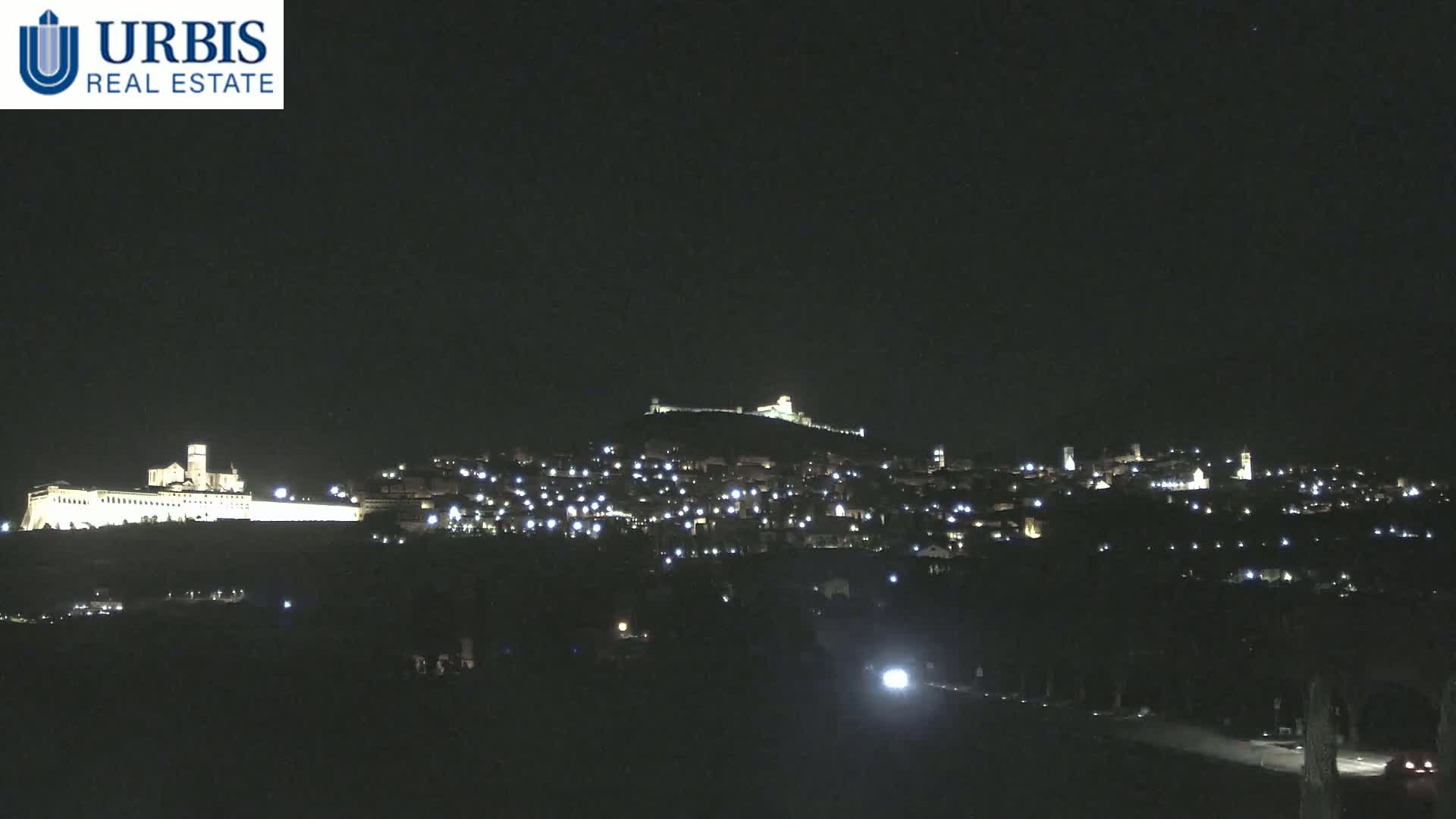 A nighttime view of a hilltop town illuminated by lights, with a large building complex also lit up on a lower hill in the background, under a clear, dark sky.