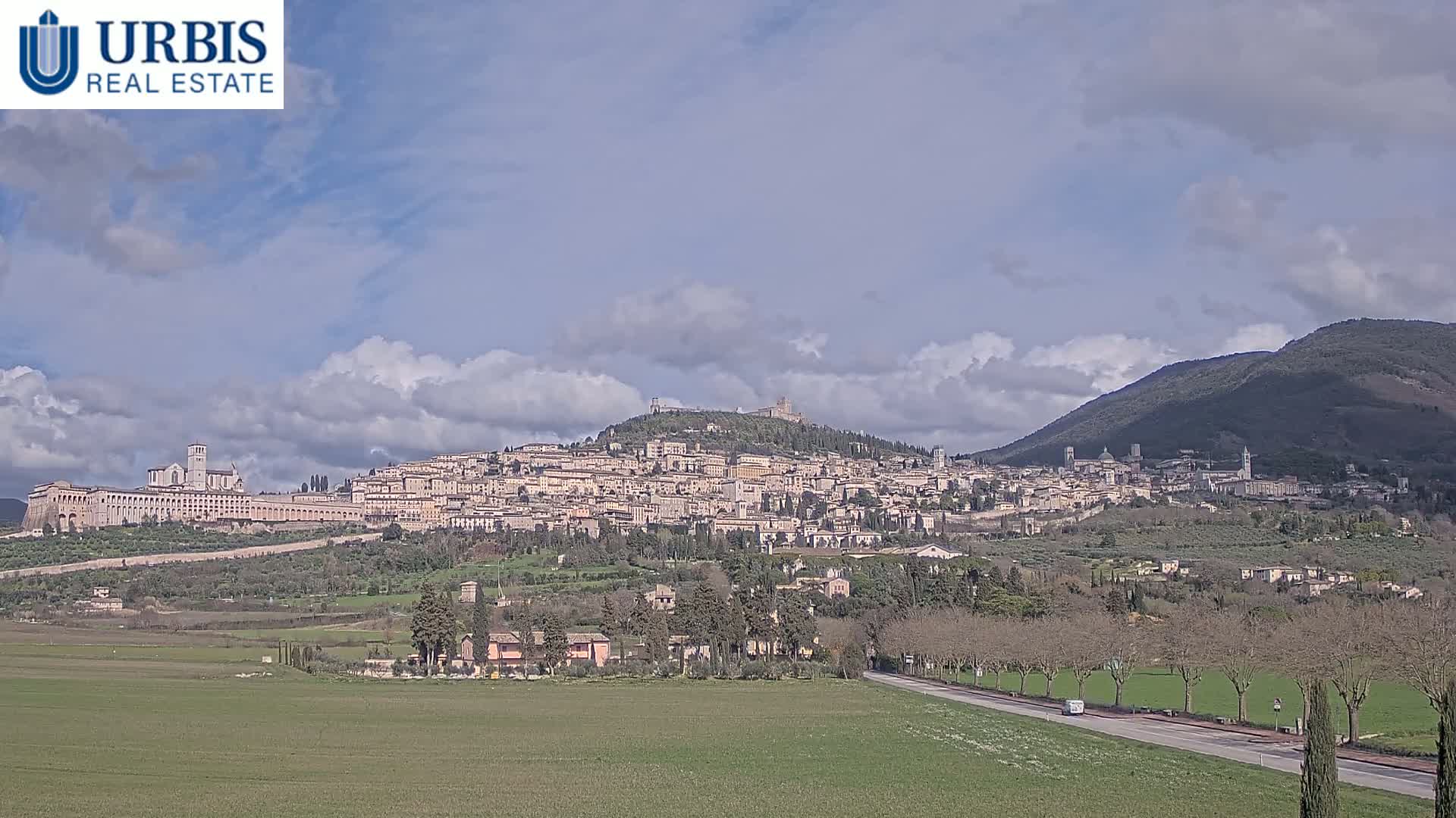 A large hill town sits under a partly cloudy sky, with green fields and trees in the foreground.