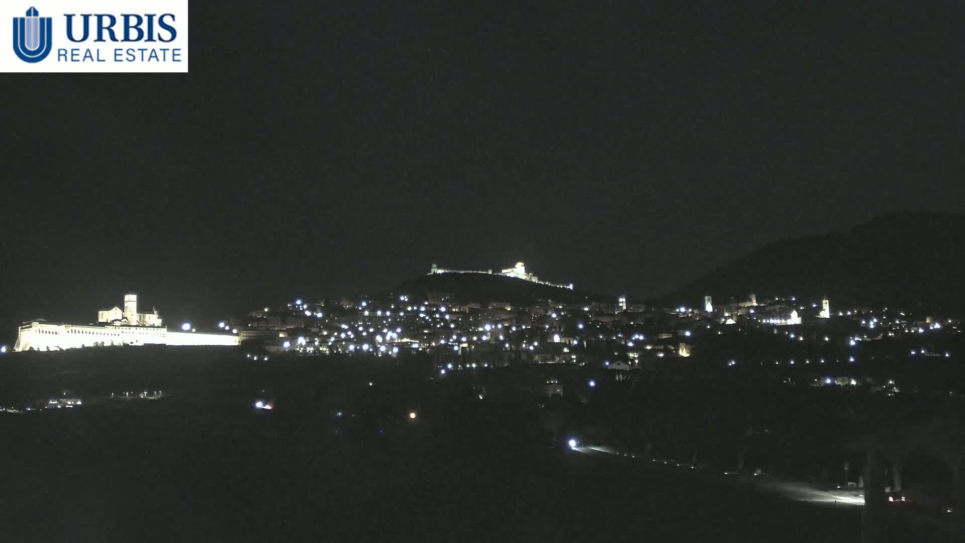 A nighttime view of a hilltop town, illuminated by lights, with a large, brightly lit building on a lower hill in the background.