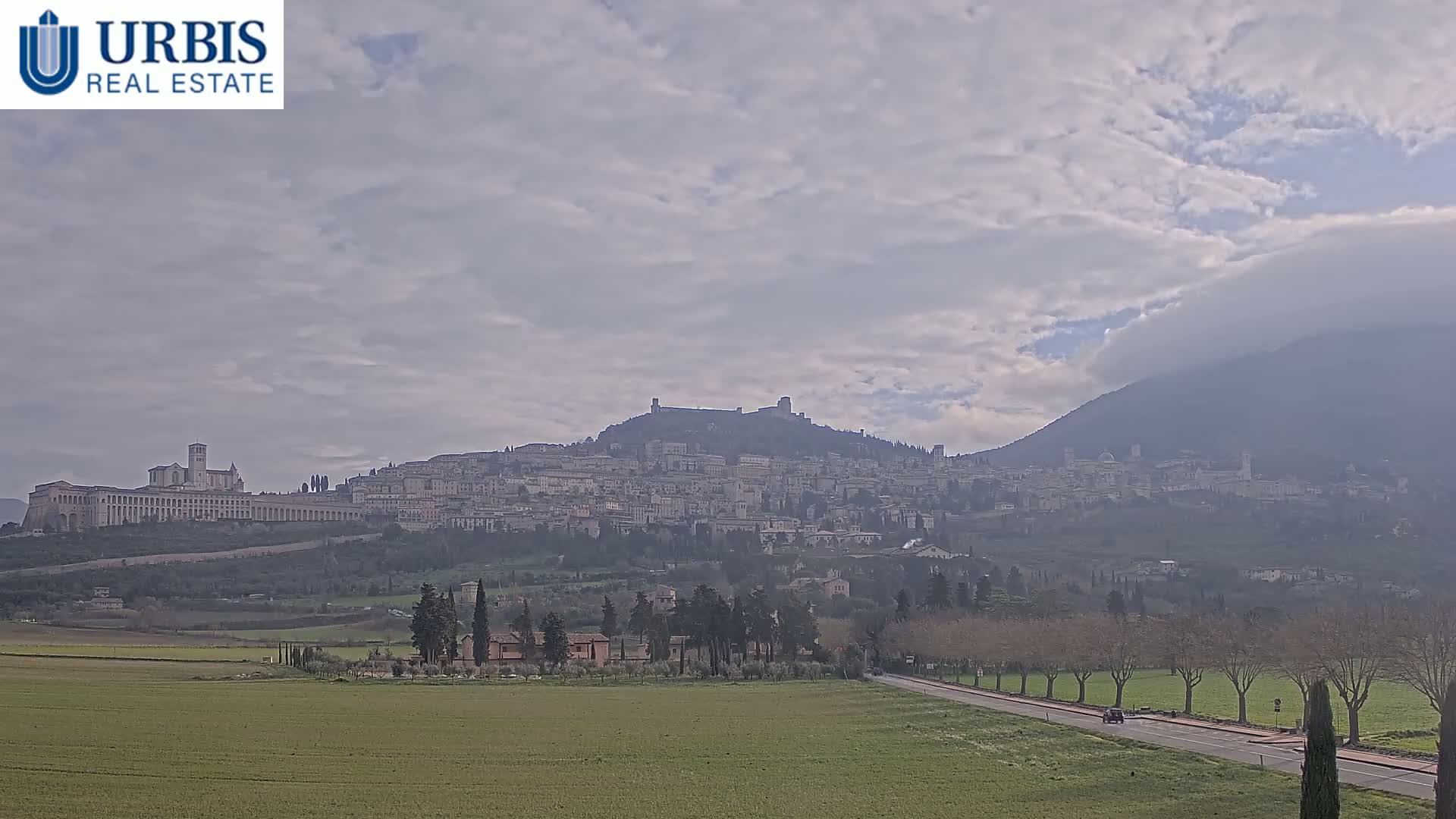 A hilltop town with a large basilica stretches across a hillside under a mostly cloudy sky, with a flat, green field and road in the foreground.