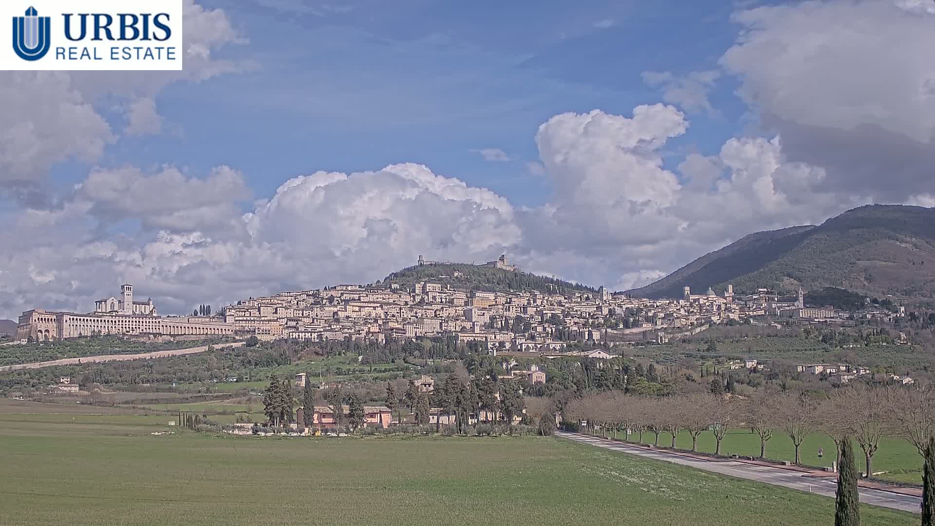 A hilltop town with many buildings, under a partly cloudy sky, is seen from a distance across a flat green field and a road.