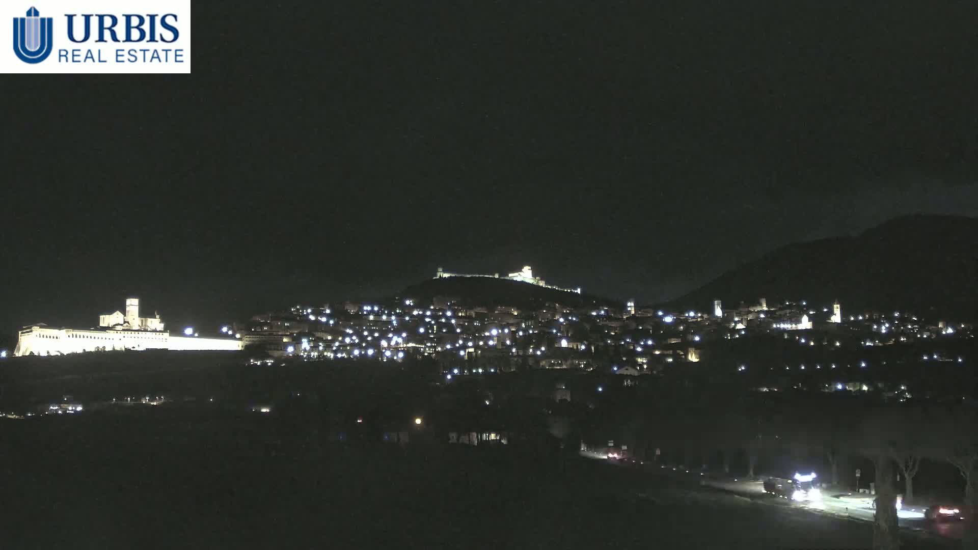 A nighttime view of a hilltop town illuminated by lights, with a larger structure, possibly a church or monastery, prominently lit at the base of the hill.