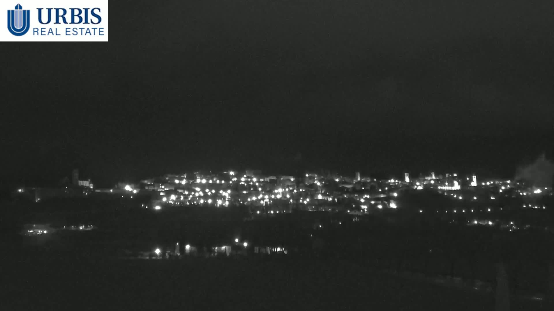 A nighttime long shot shows a small town's lights spread across a hill under a dark, cloudy sky.