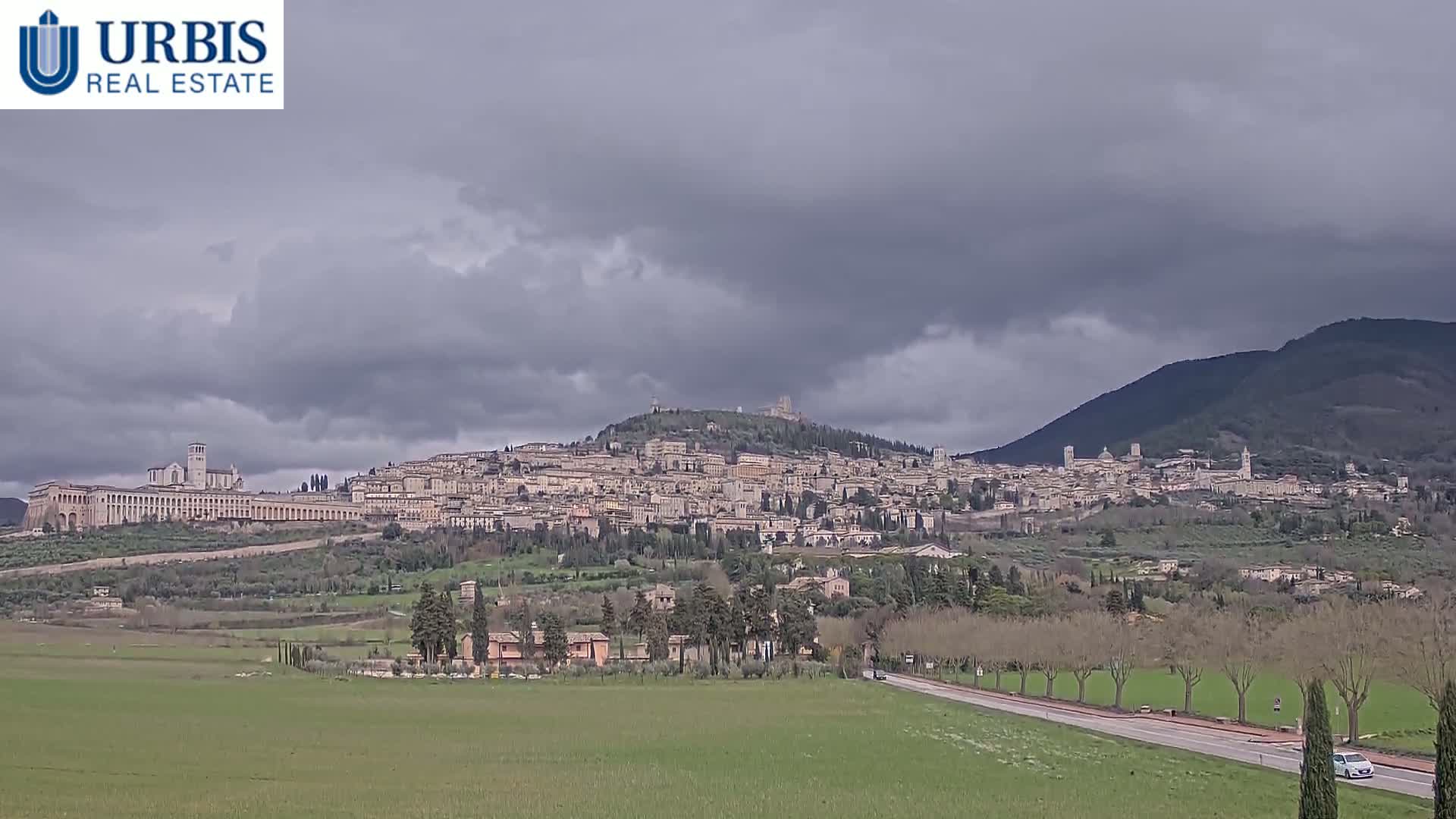 A sprawling hilltop town, under a cloudy sky, is situated in a verdant valley alongside a road and adjacent to a mountain.