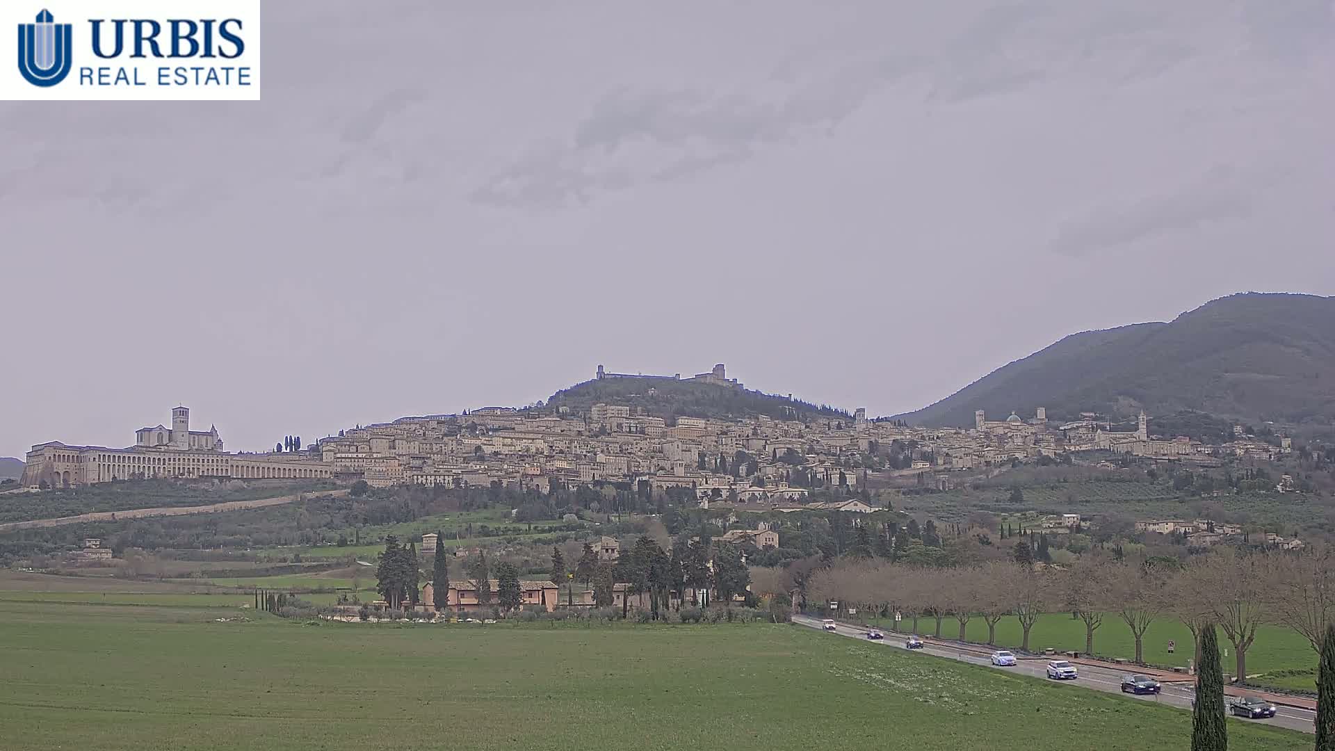 A hilltop town with many buildings and a large church is visible on a cloudy day, with a flat green field in the foreground and a road with cars in the middle ground.