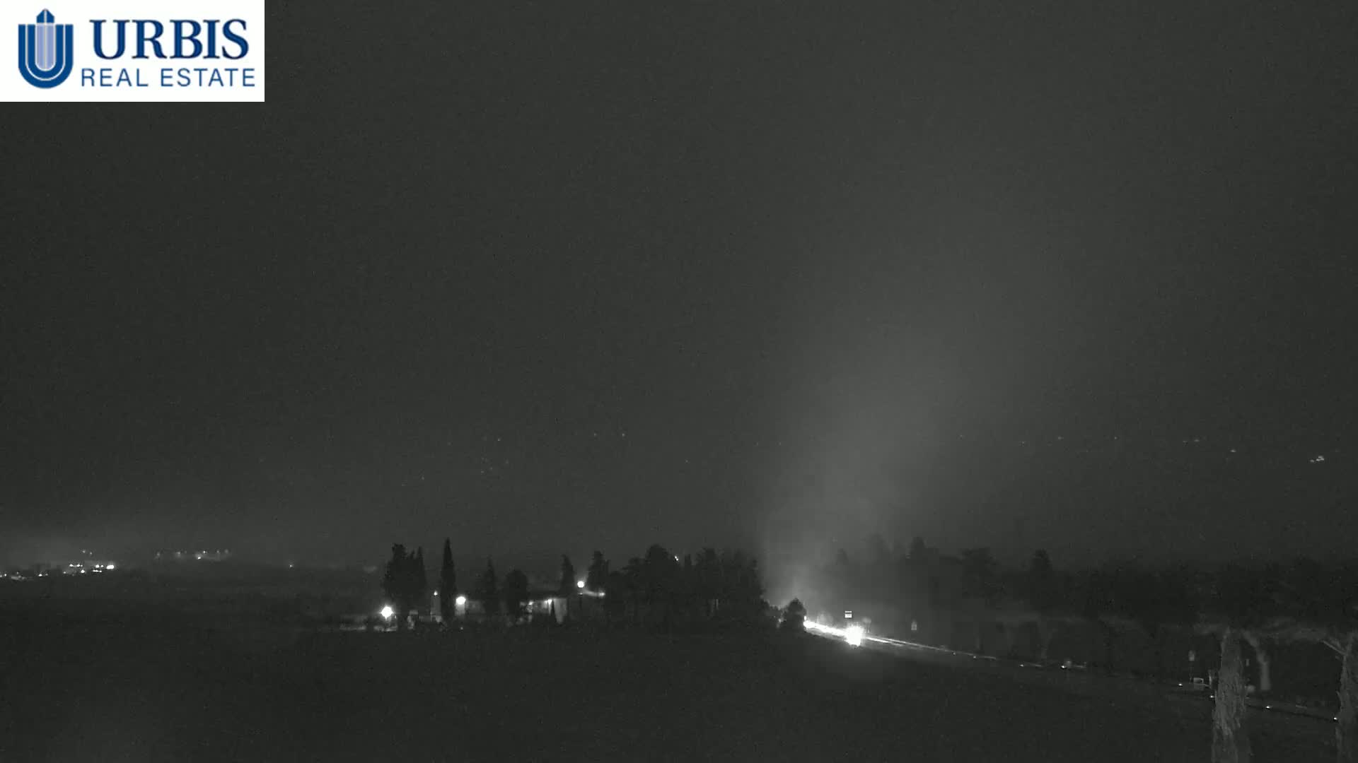 A nighttime, hazy view of a road with vehicles driving through a landscape featuring sparsely distributed trees and distant lights.