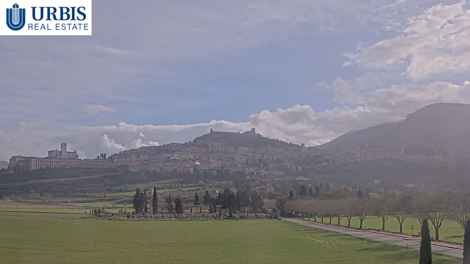 A hilltop town, partially obscured by haze, sits beneath a partly cloudy sky, with a large flat expanse of green field in the foreground.