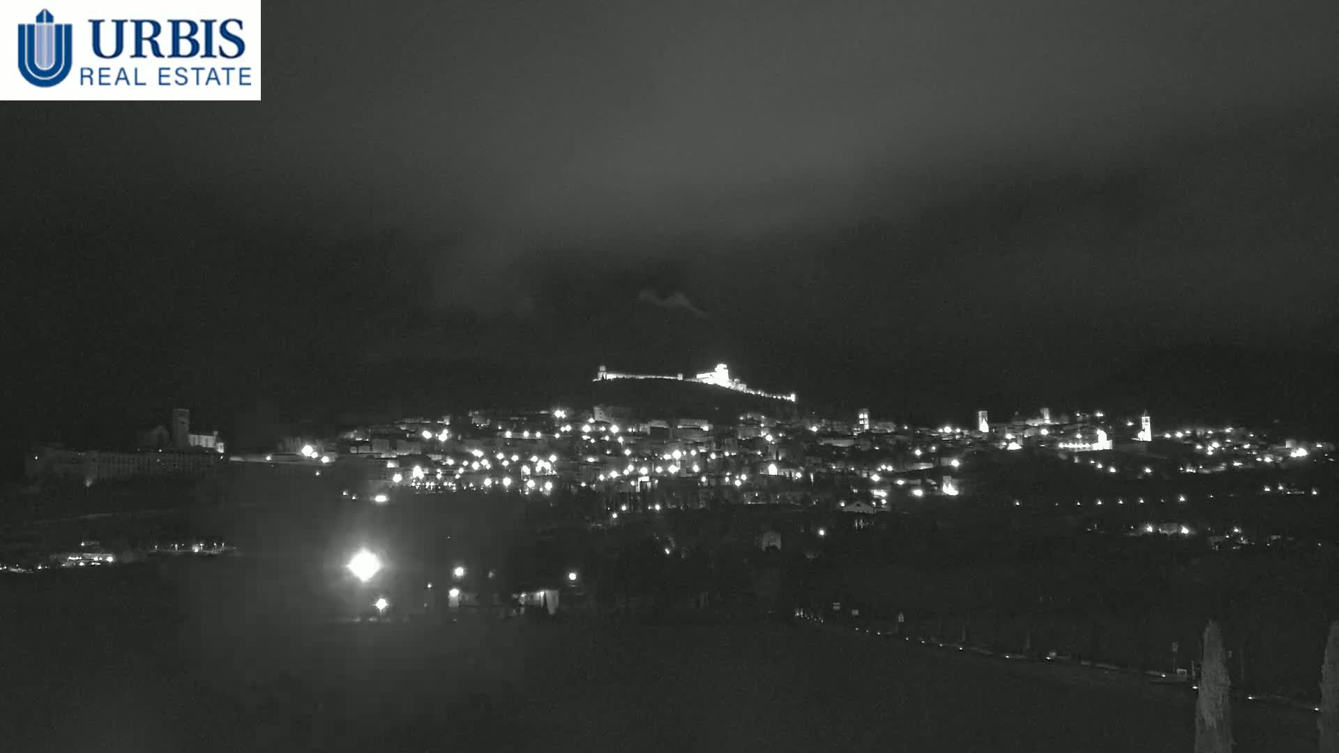 A nighttime, long shot of a town nestled on a hill, illuminated by scattered lights under a dark, cloudy sky.
